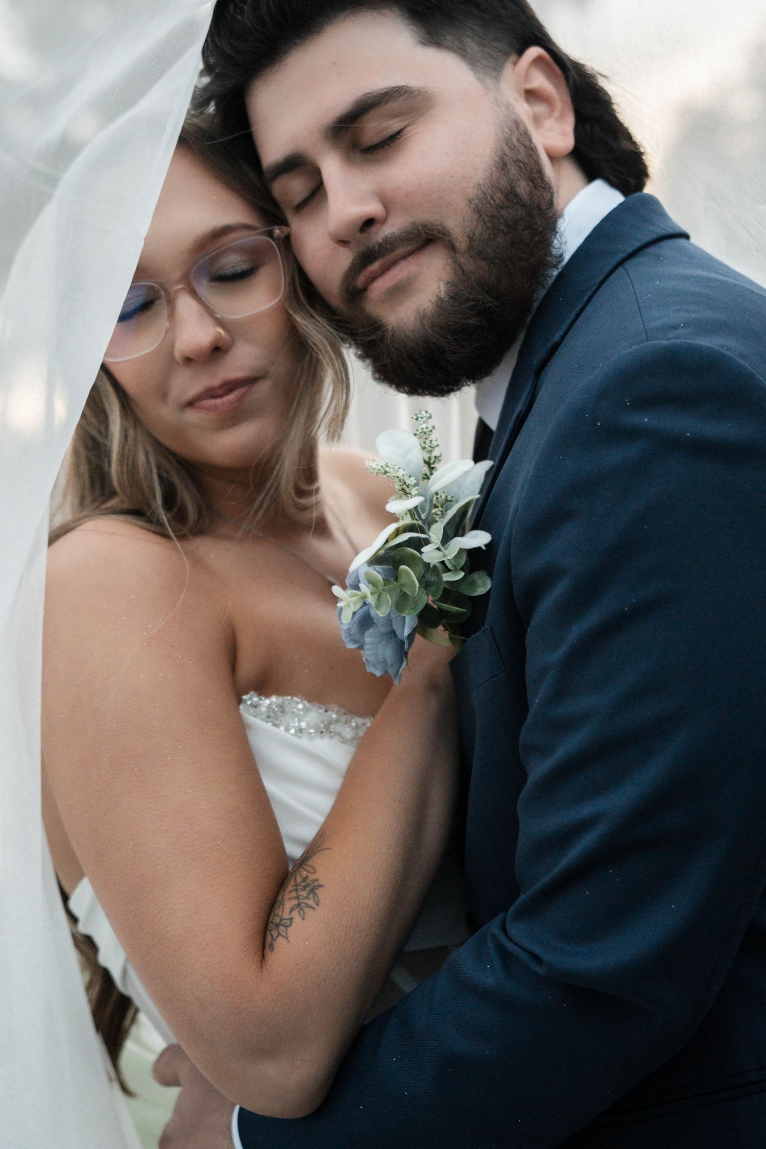 Groom tenderly embraces bride under sheer veil, both with closed eyes in intimate wedding portrait.