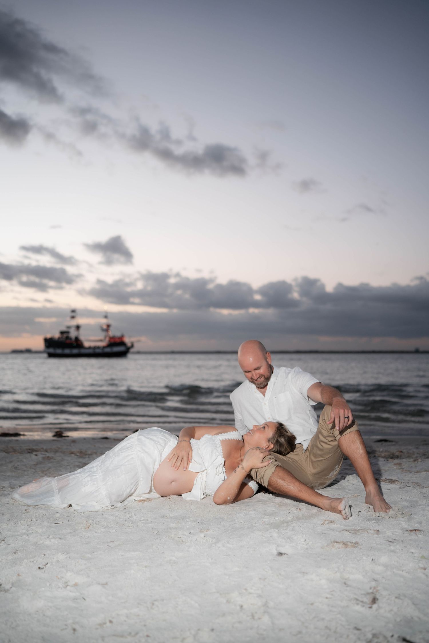 Bald man tenderly gazing at pregnant partner lying on beach sand at dusk with boat in background.