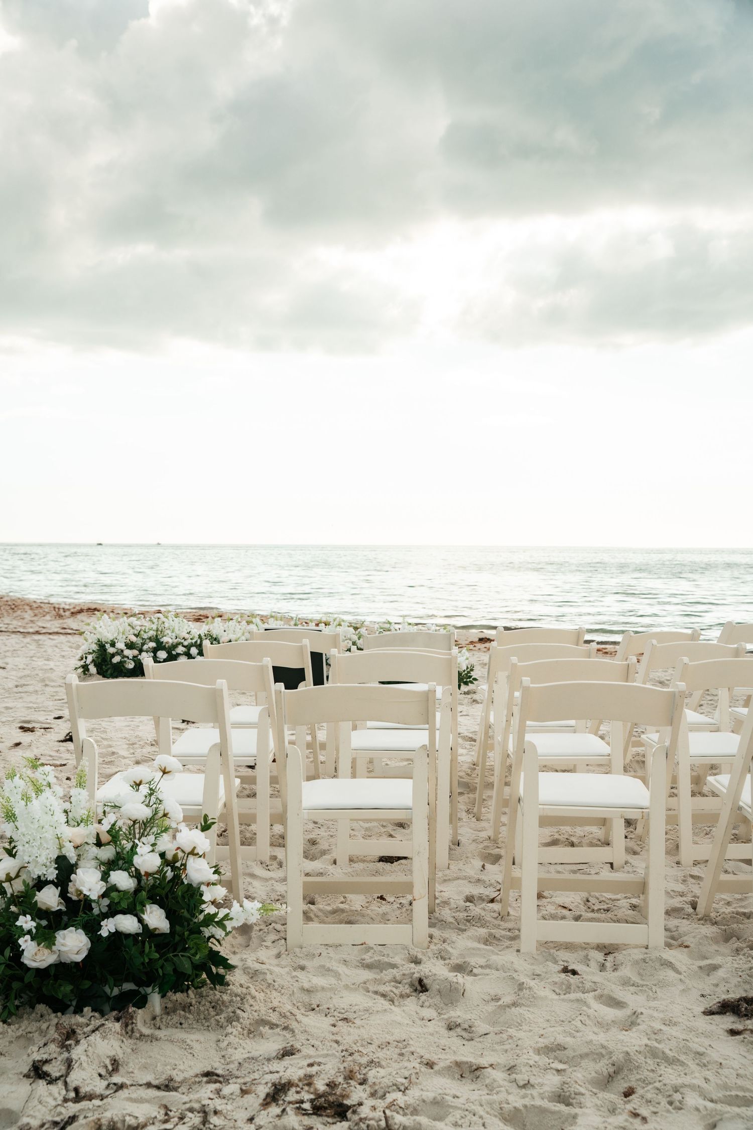 White folding chairs arranged on sandy beach facing ocean under dramatic cloudy sky for wedding.
