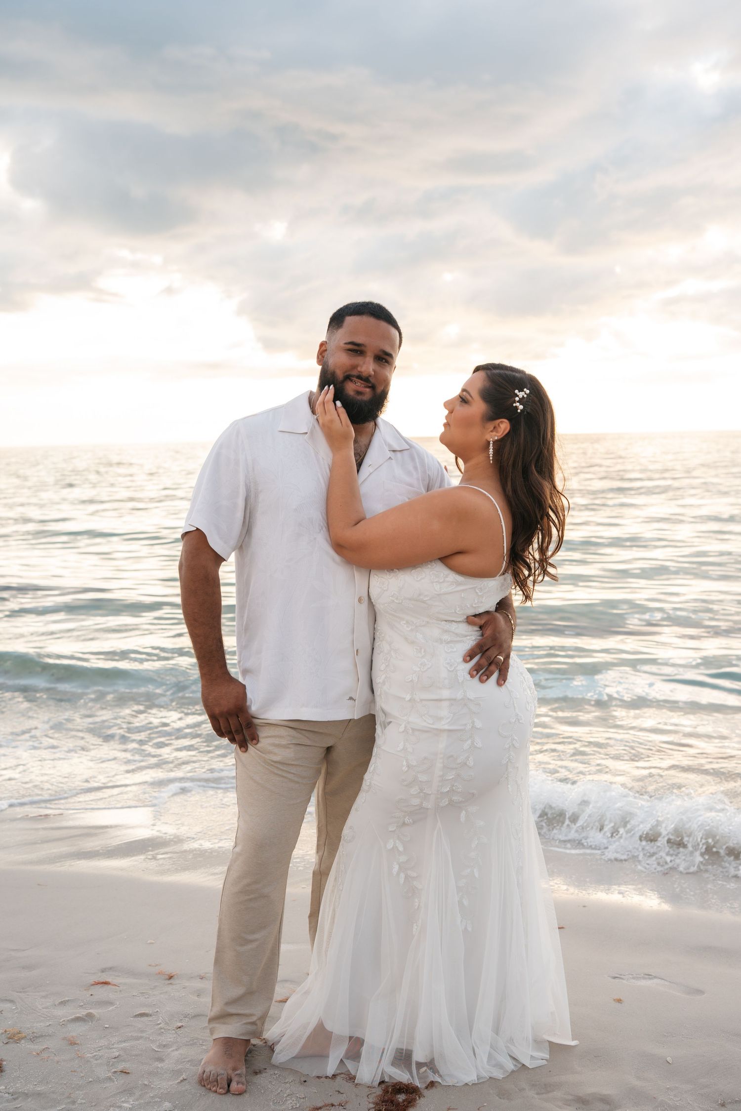Newlywed couple posing together on beach at sunset, bride in lace gown.