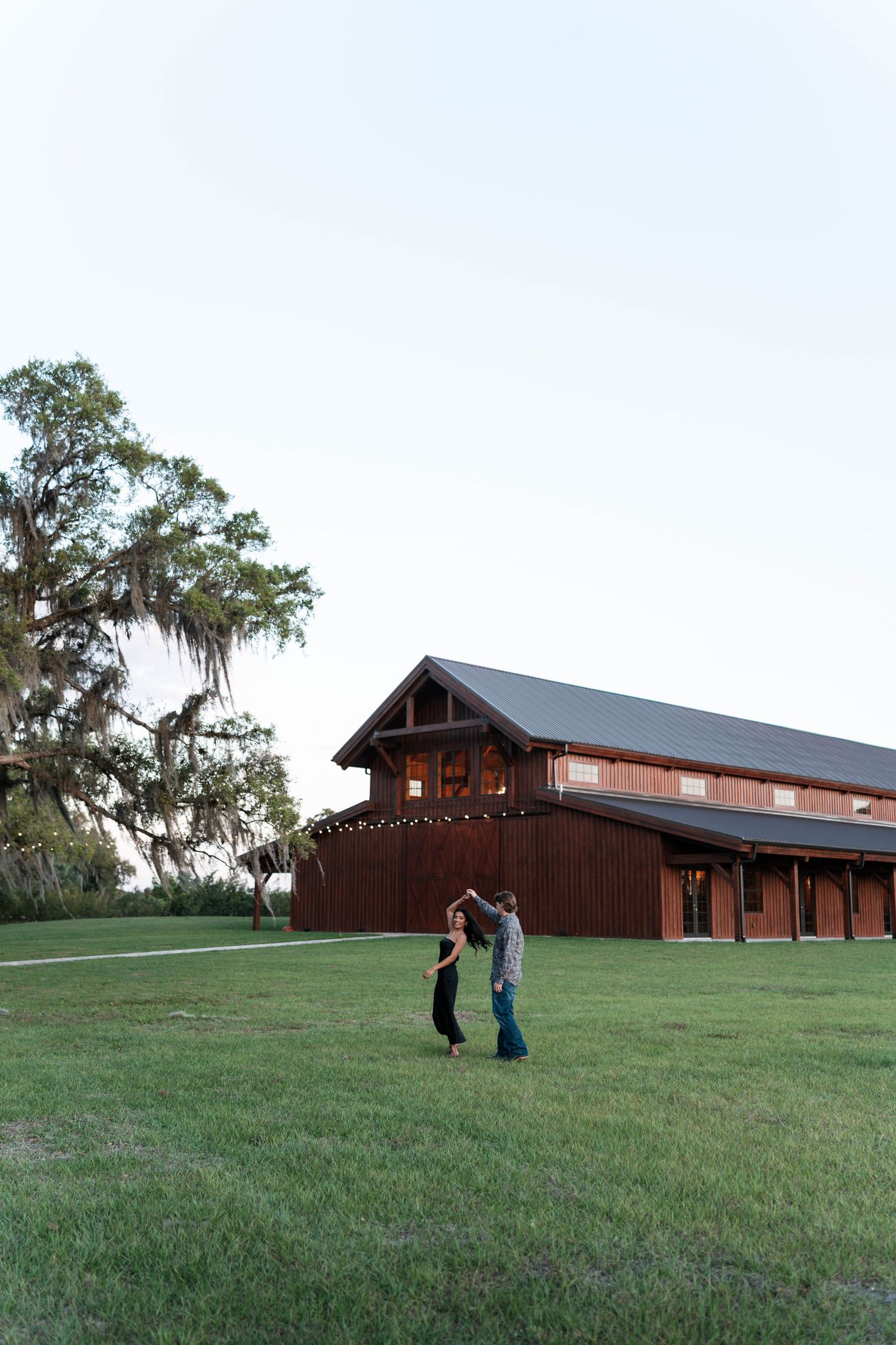 Couple dancing on green lawn outside rustic brown barn wedding venue with Spanish moss oak tree at sunset.