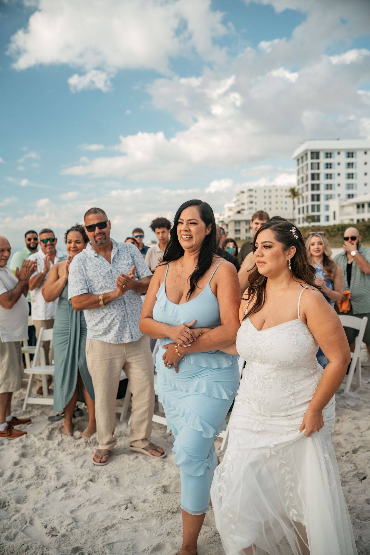 Bride walking down beach aisle with bridesmaid in blue dress, guests applauding.