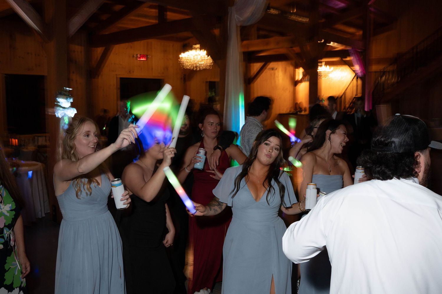 Bridesmaids in dusty blue dresses dance with glow sticks at barn wedding reception.