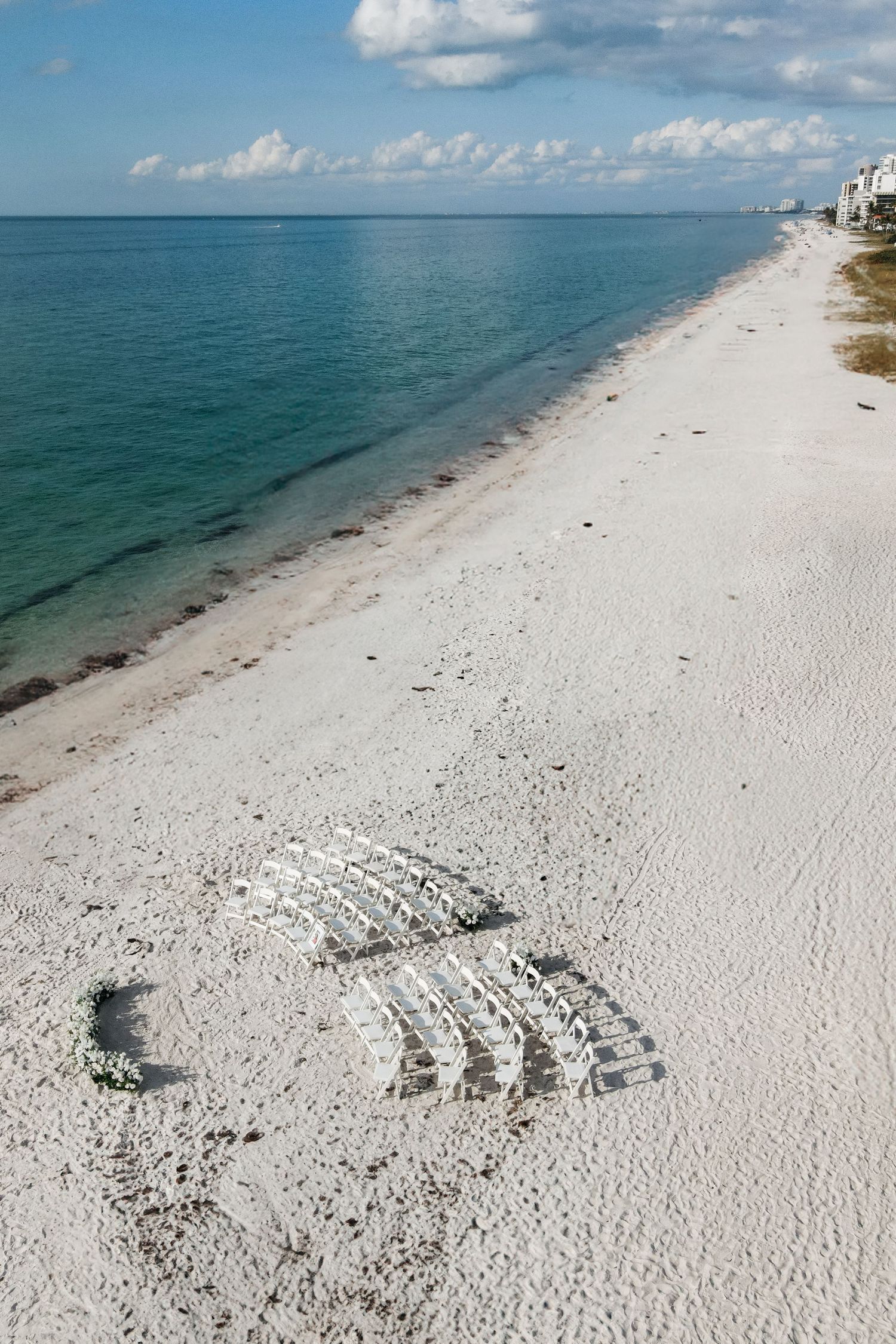 Aerial view of white folding chairs arranged on pristine white sandy beach for wedding ceremony.