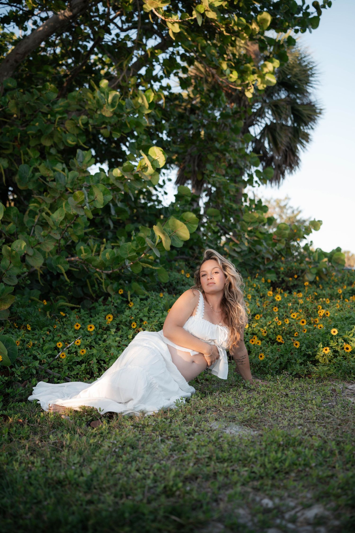 Pregnant woman in white dress sitting among wildflowers and lush greenery during golden hour outdoor maternity shoot.