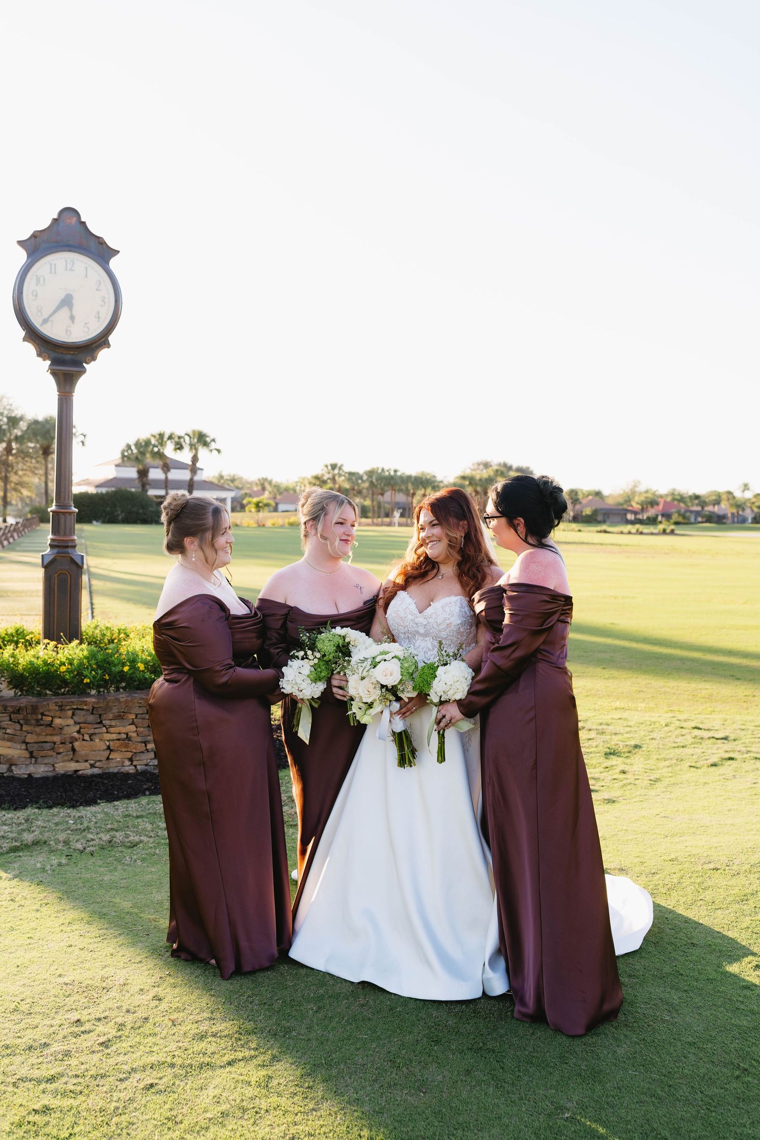 Bride with three bridesmaids in dark red off-shoulder gowns posing by a golf course clock.