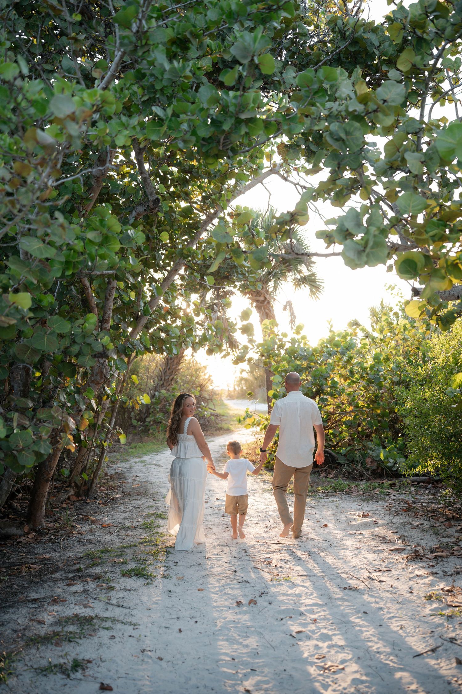 Family of three walking hand-in-hand down a sun-drenched sandy path lined with lush green trees at golden hour.