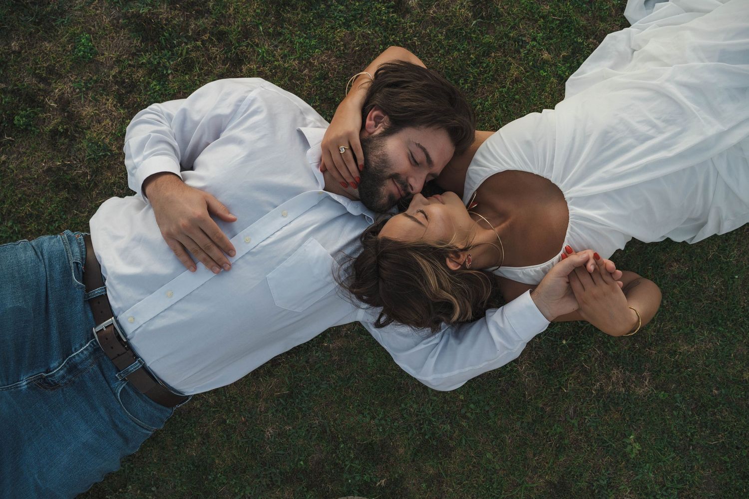 Couple in matching white shirts lie on green grass holding hands in tender aerial overhead portrait.
