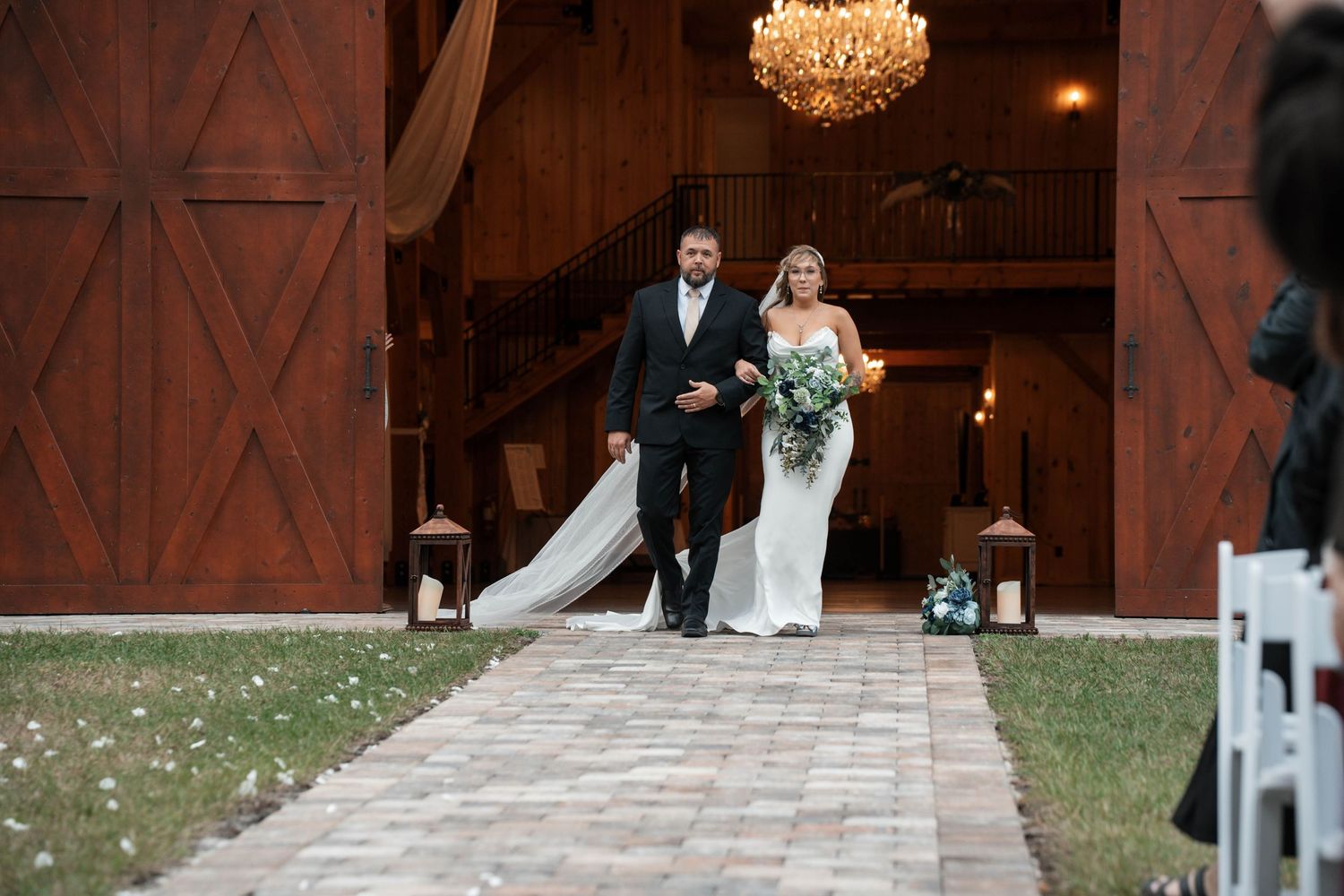 Bride in white gown walks down aisle from rustic barn with escort at outdoor wedding ceremony.