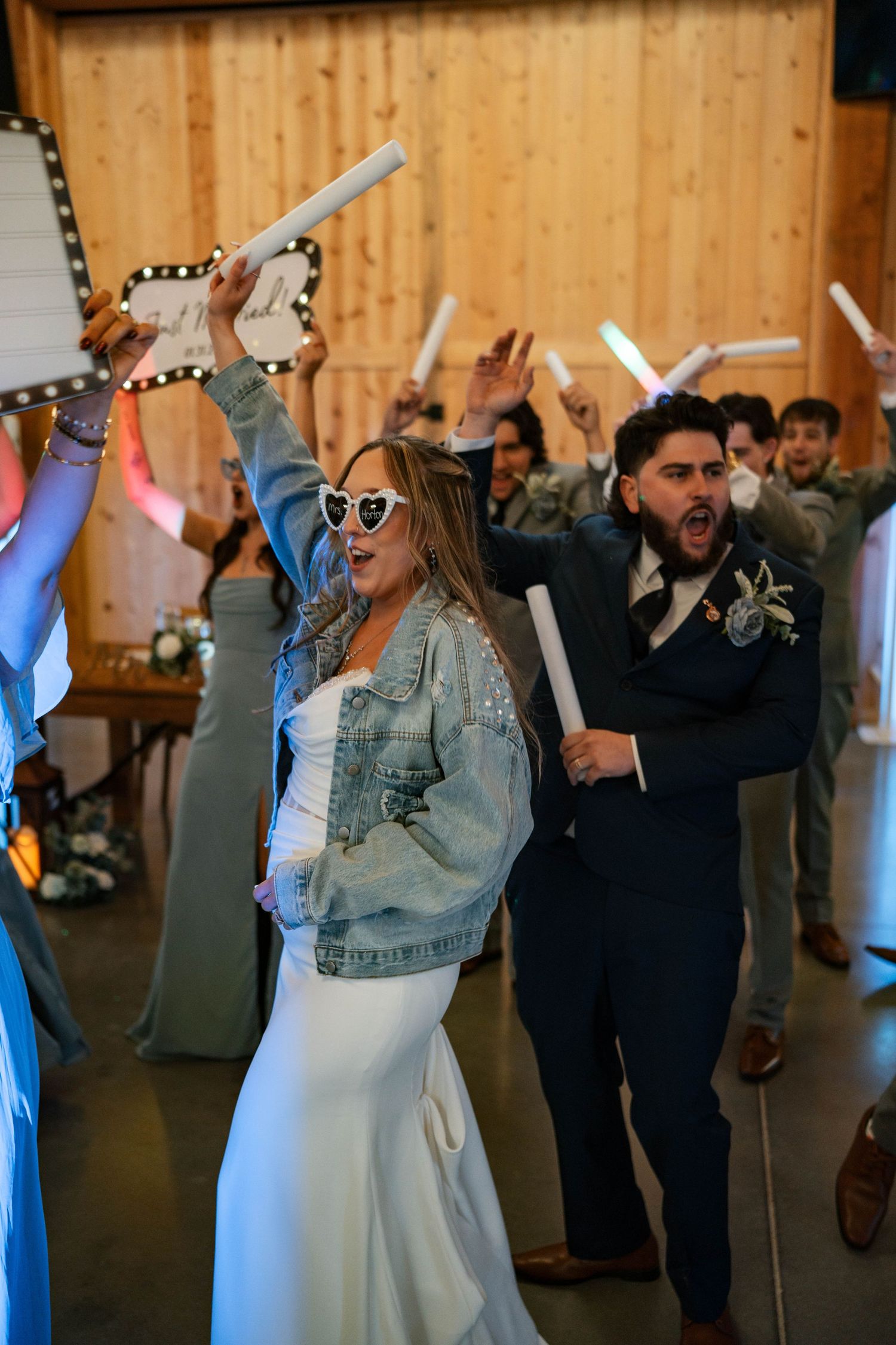 Bride in denim jacket and heart sunglasses celebrates with groom at wedding reception.