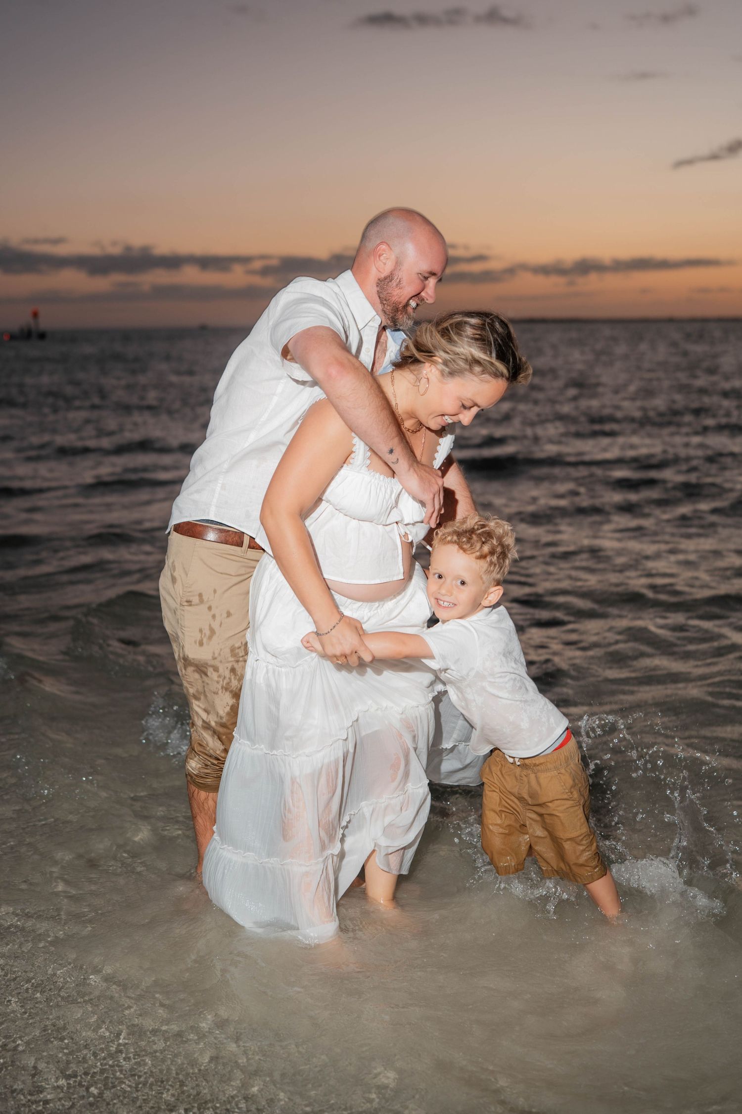 Family of three in white outfits playing in ocean waves at sunset beach, pregnant mother with husband and young son.