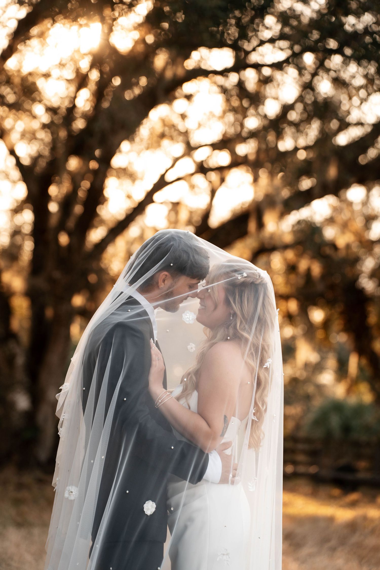 Bride and groom share an intimate moment under a pearl-adorned veil in golden sunlit forest.
