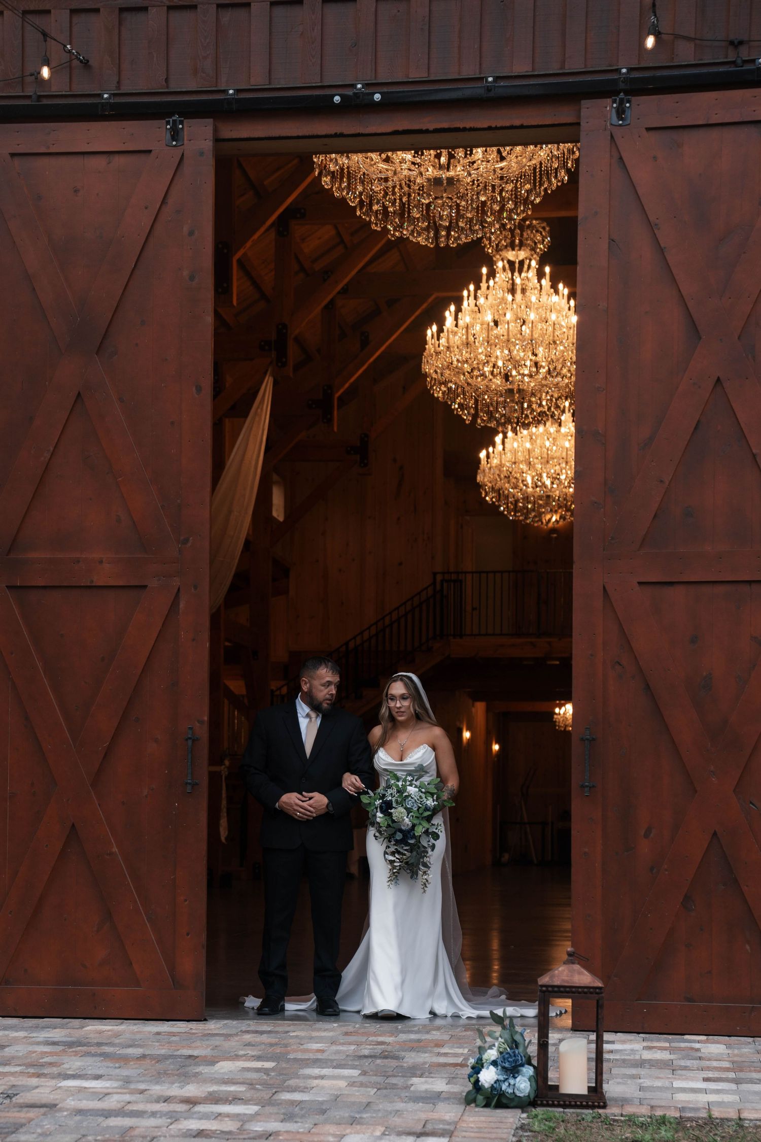 Bride and escort stand in grand barn venue doorway framed by stunning crystal chandeliers.