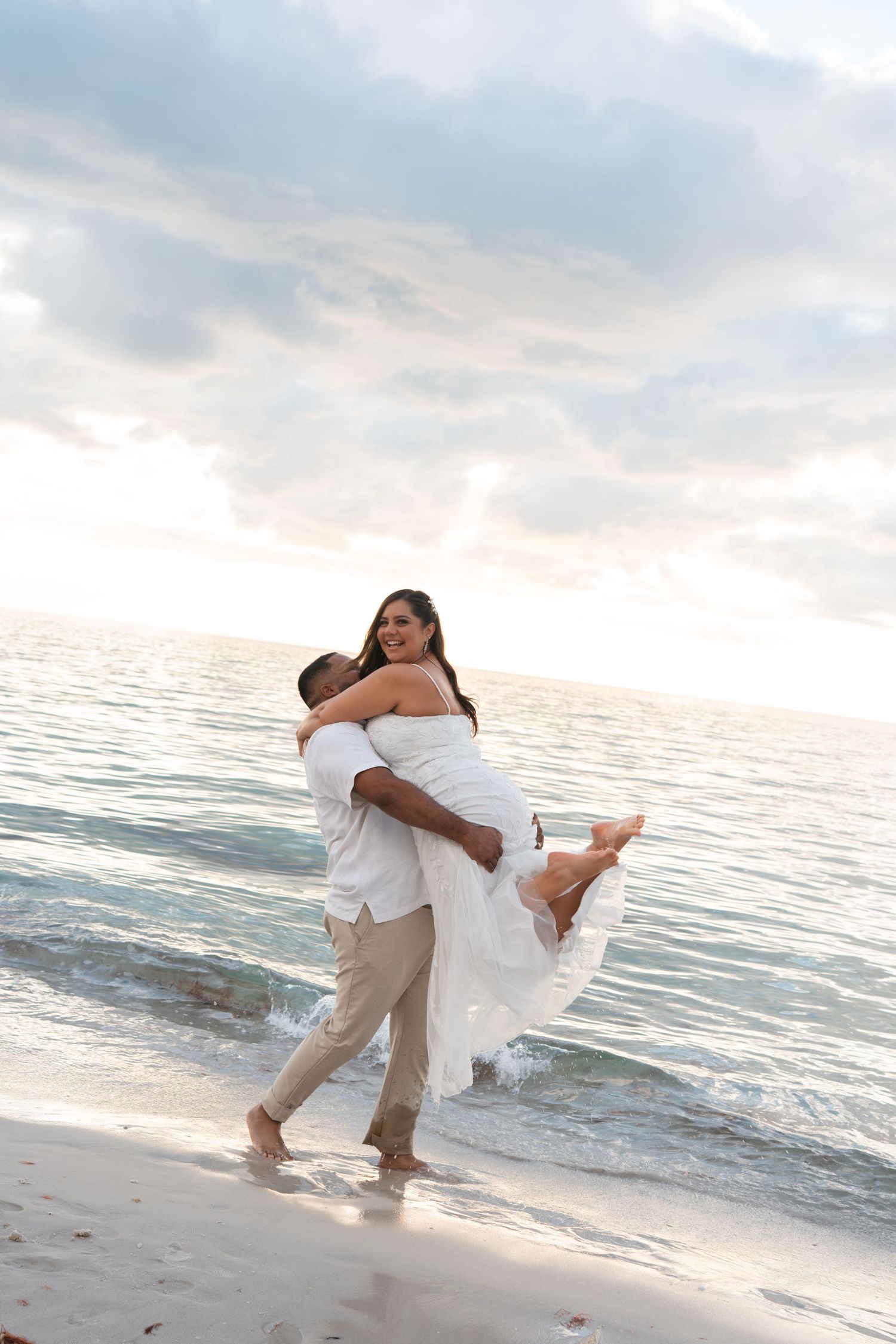 Groom lifting smiling bride in white dress on sandy beach at sunset.