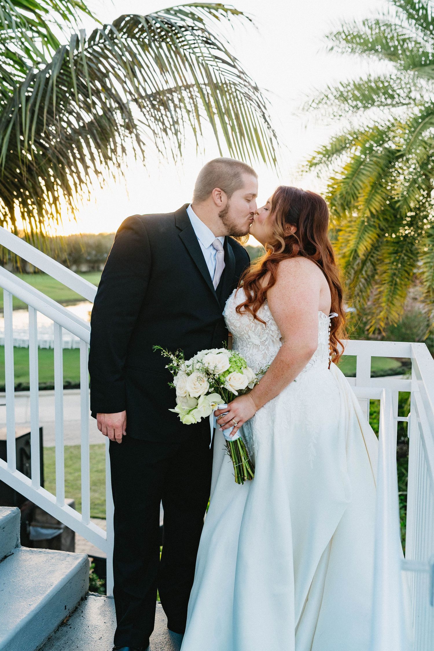 Bride and groom share a romantic kiss on white staircase surrounded by palm trees at golden hour.