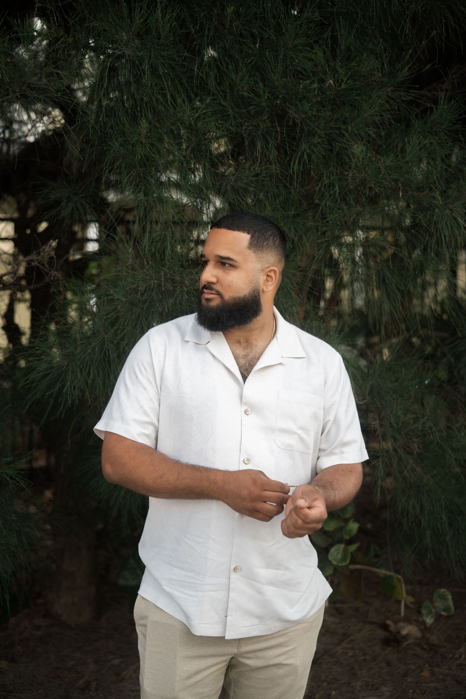 Bearded man in white linen shirt stands confidently in front of lush green pine trees.