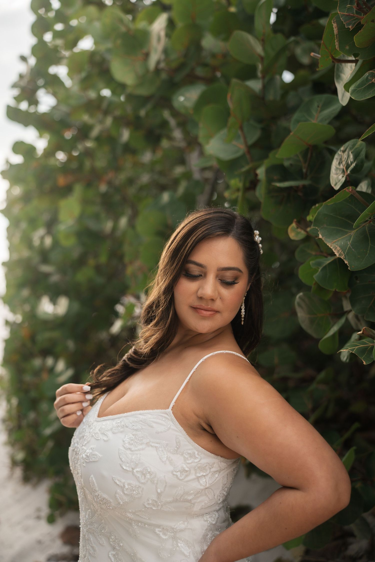 Beautiful bride in lace wedding gown poses between lush green tropical foliage on beach.