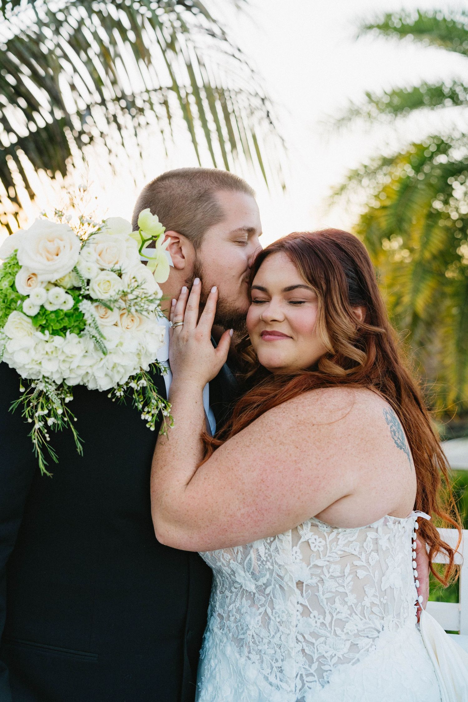 Groom tenderly kisses bride on cheek as she smiles holding white floral bouquet outdoors.