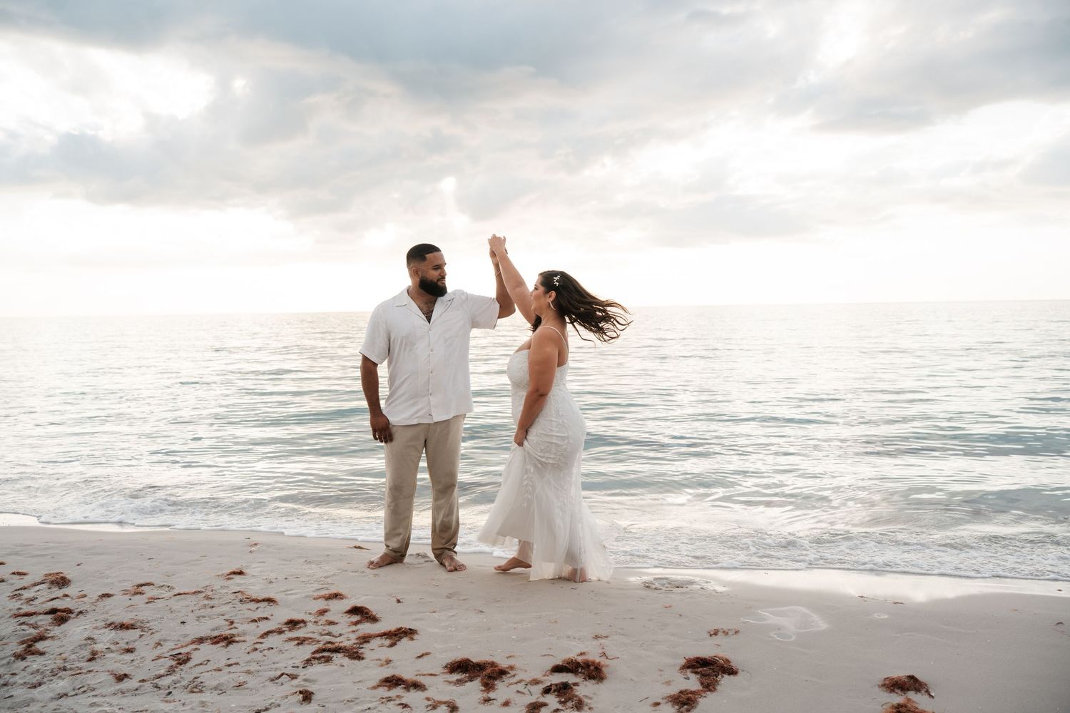 Couple in white attire dancing on a sandy beach at sunset with calm ocean waves in the background.
