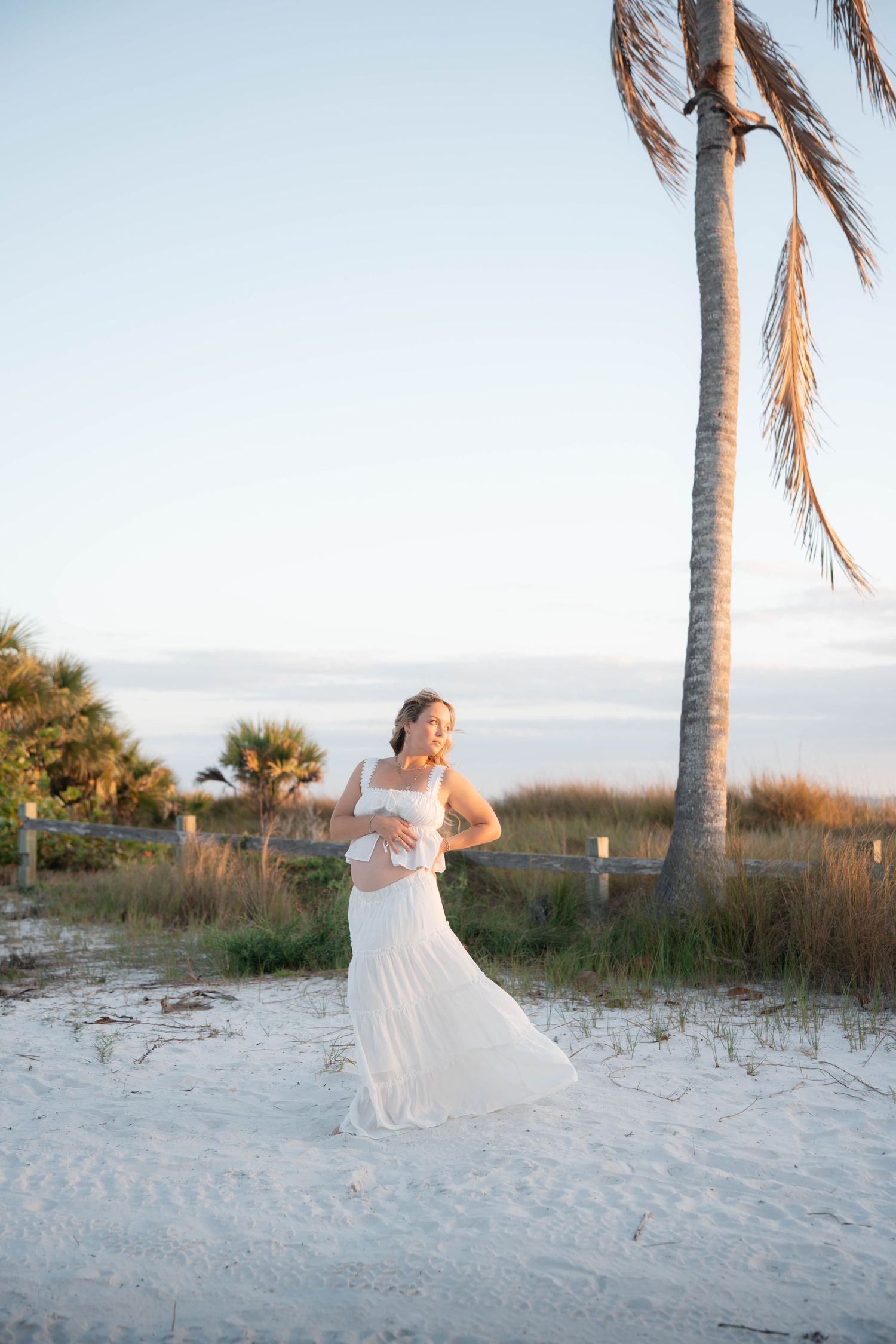 Pregnant woman in white dress standing on sandy beach near palm tree at sunset.
