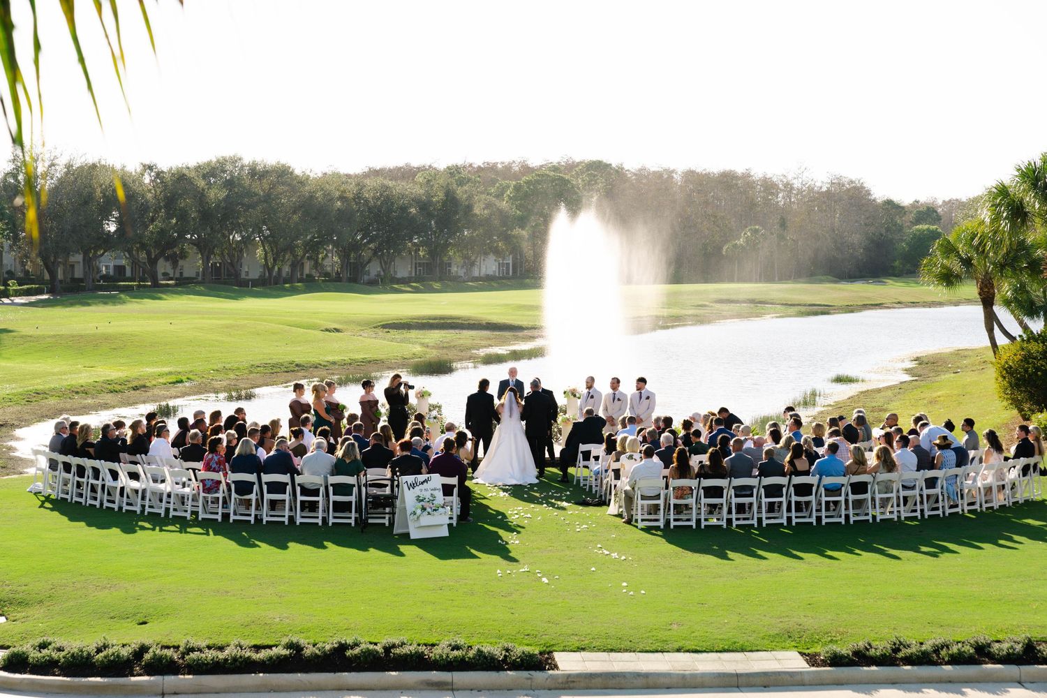 Wide shot of elegant outdoor wedding ceremony on golf course with fountain backdrop.