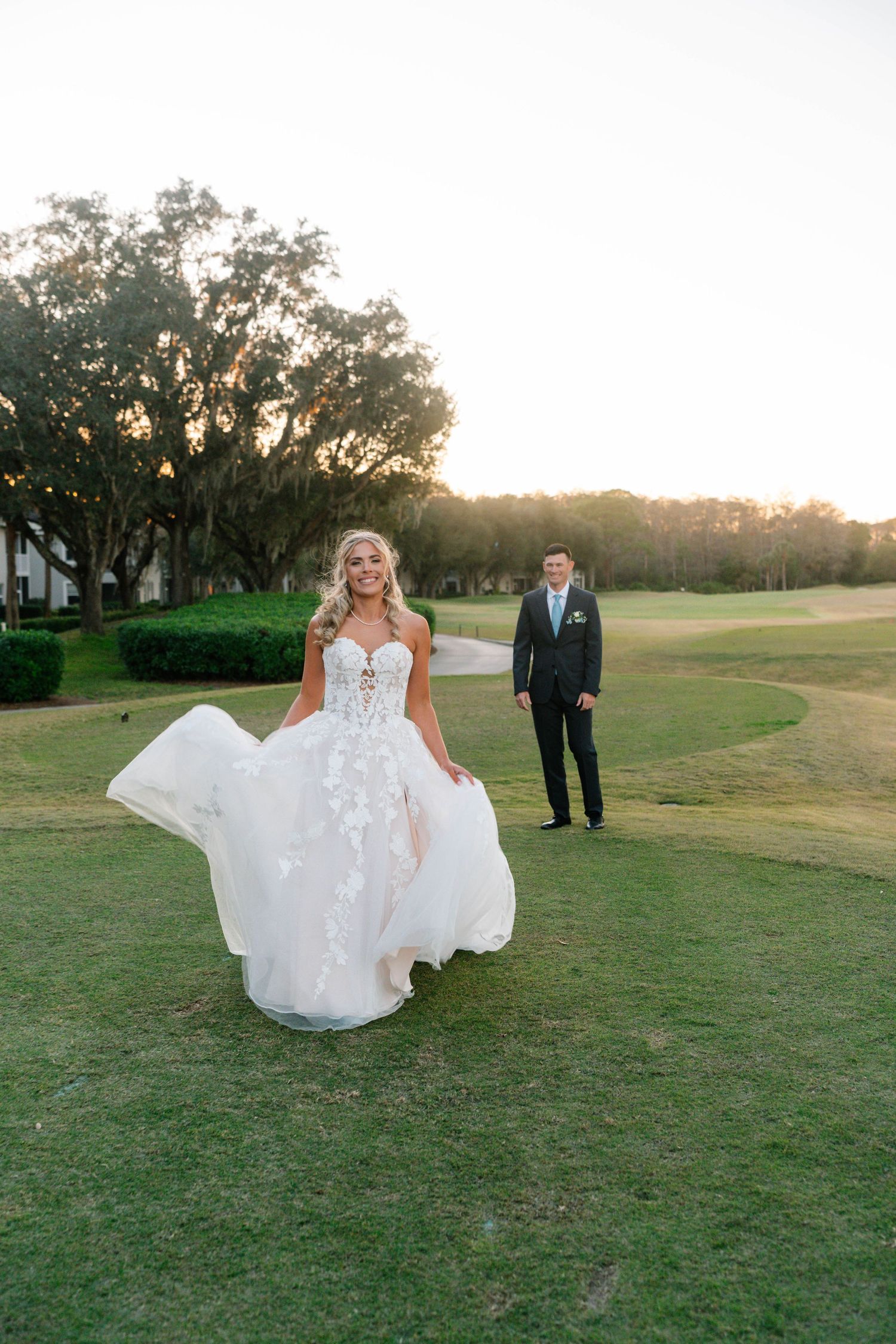 Bride in a floral ball gown twirling on a golf course at sunset with groom standing in the background.