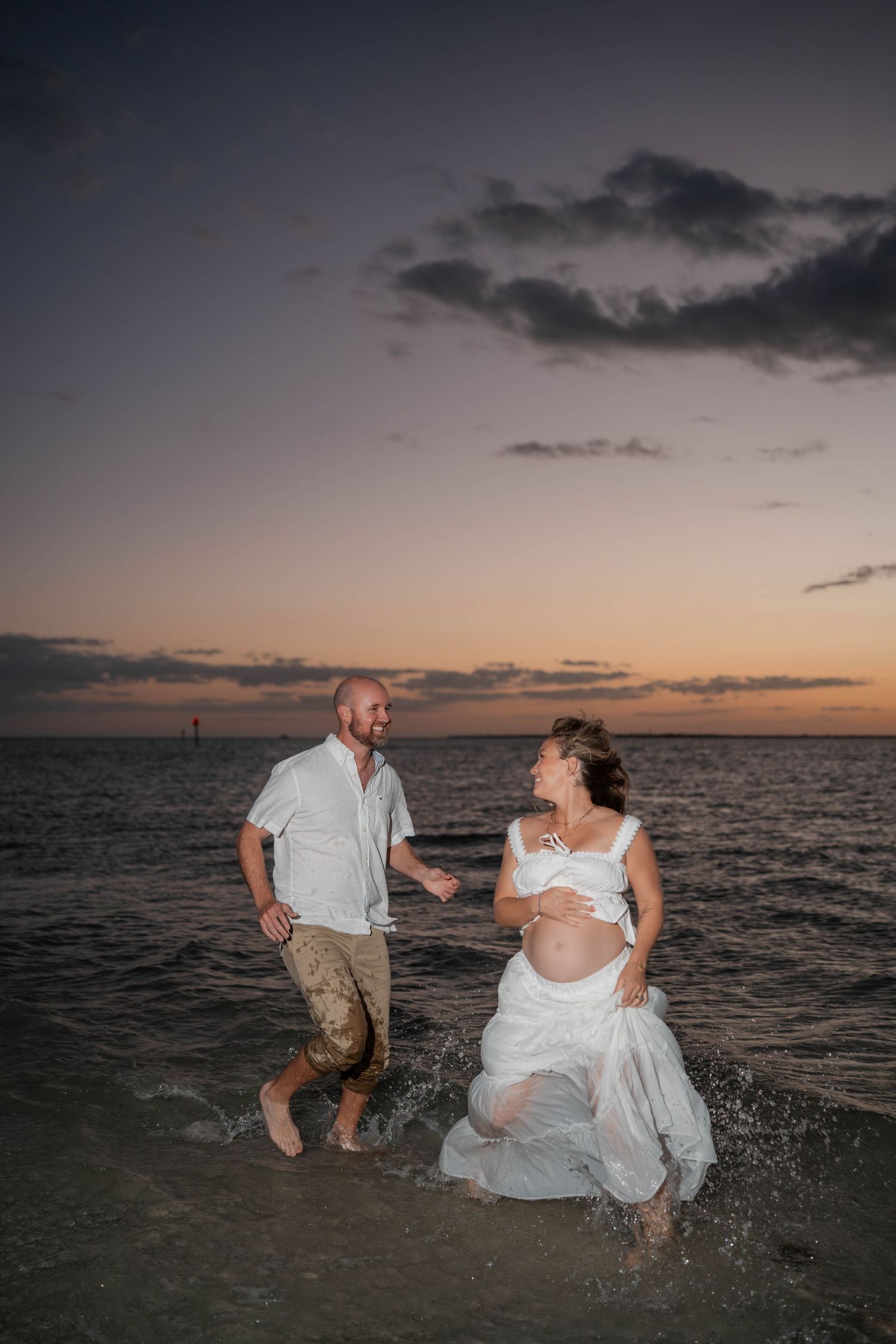 Joyful expecting couple splashing through ocean waves at colorful sunset during beach maternity photography session.