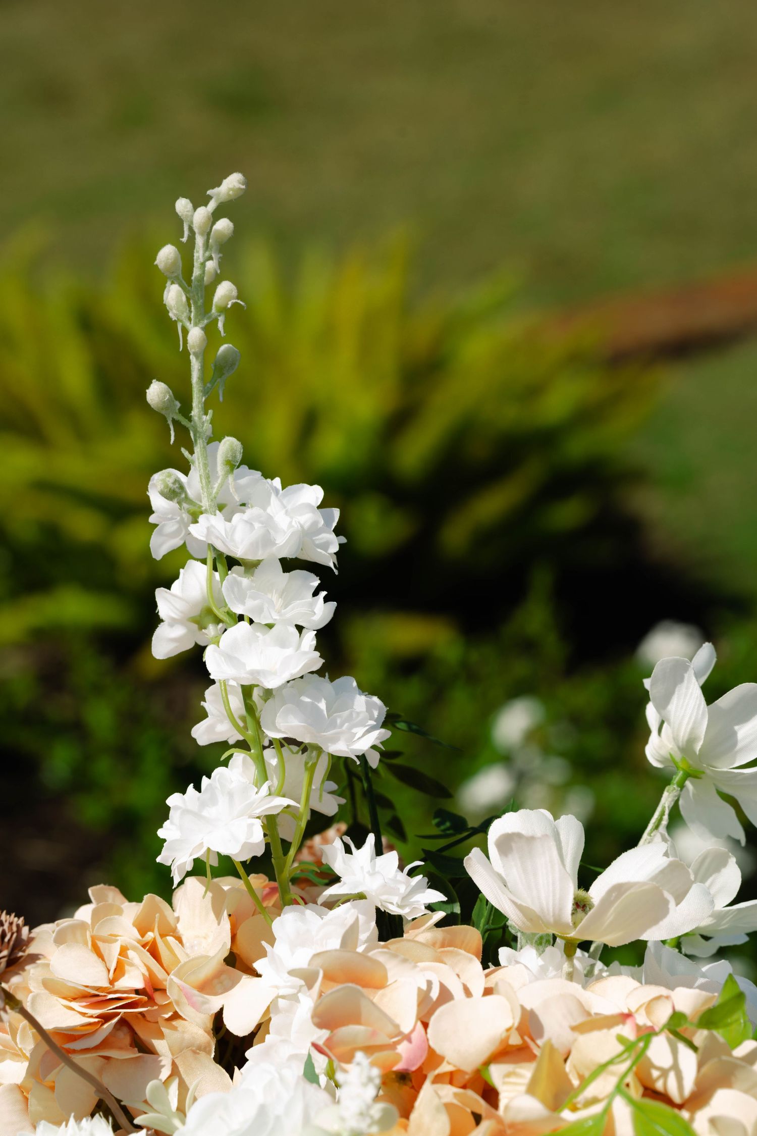 Close-up of white delphinium and peach floral wedding arrangement in a sunlit garden setting.