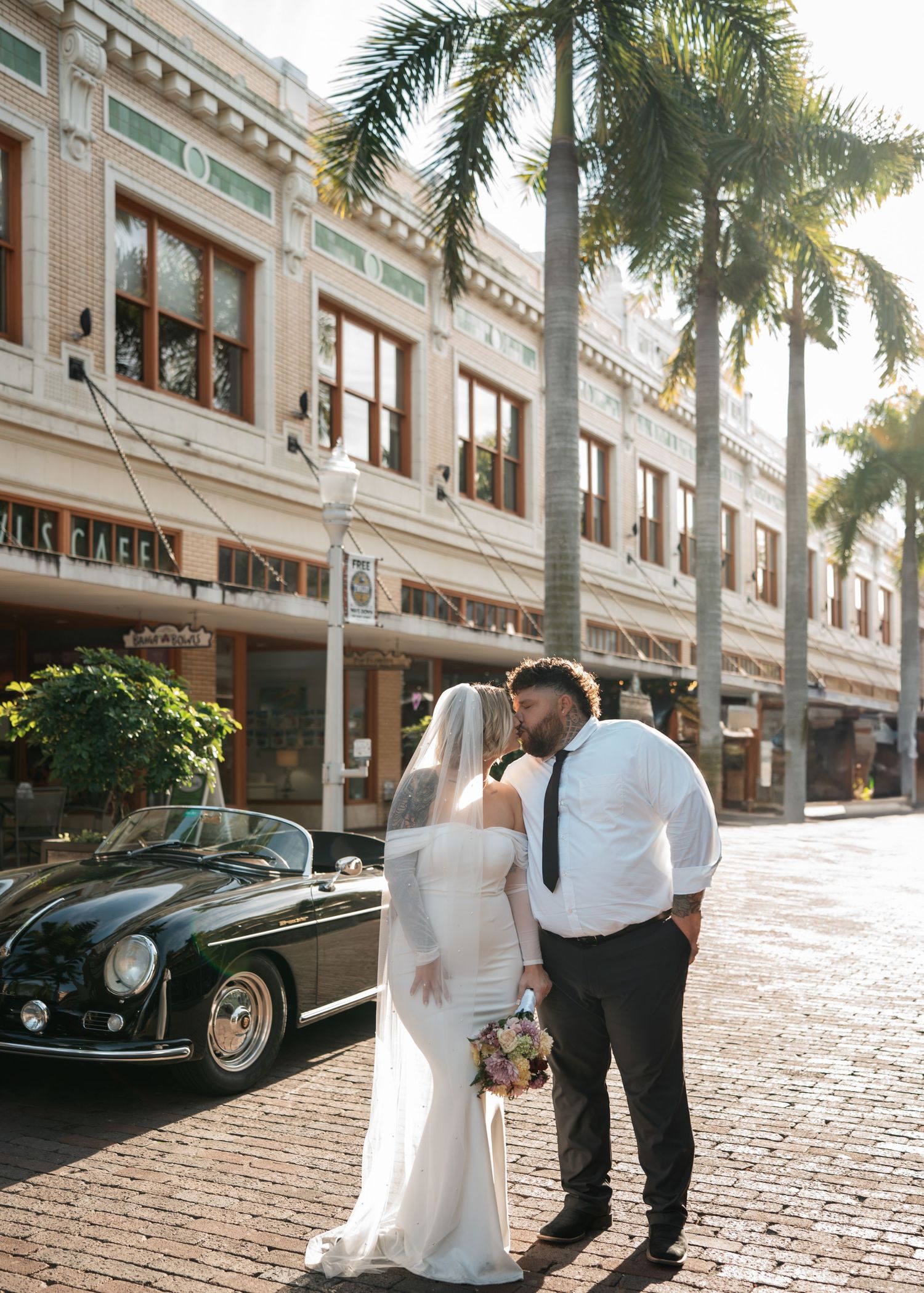 Bride and groom share a kiss on a sunny street beside a vintage black Porsche and palm trees.