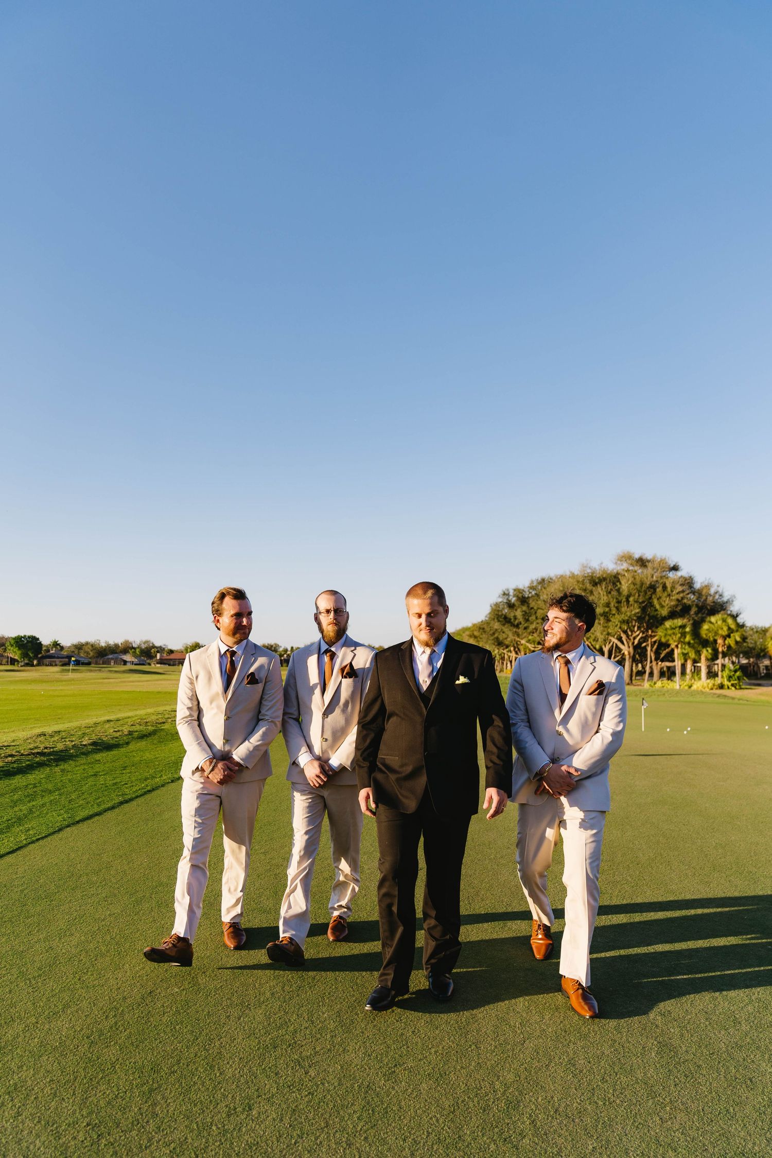 Groom and three groomsmen walk confidently across lush golf course under clear blue sky.
