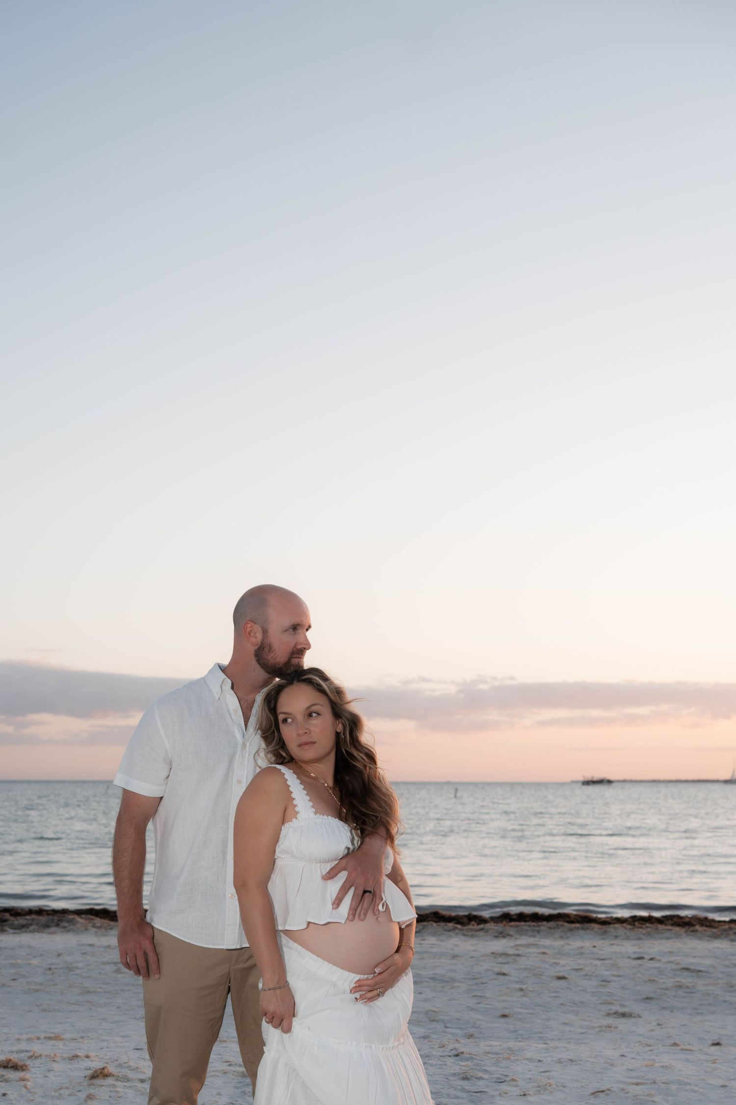 Expecting couple in white outfits embracing on sandy beach at sunset during romantic maternity photo session.