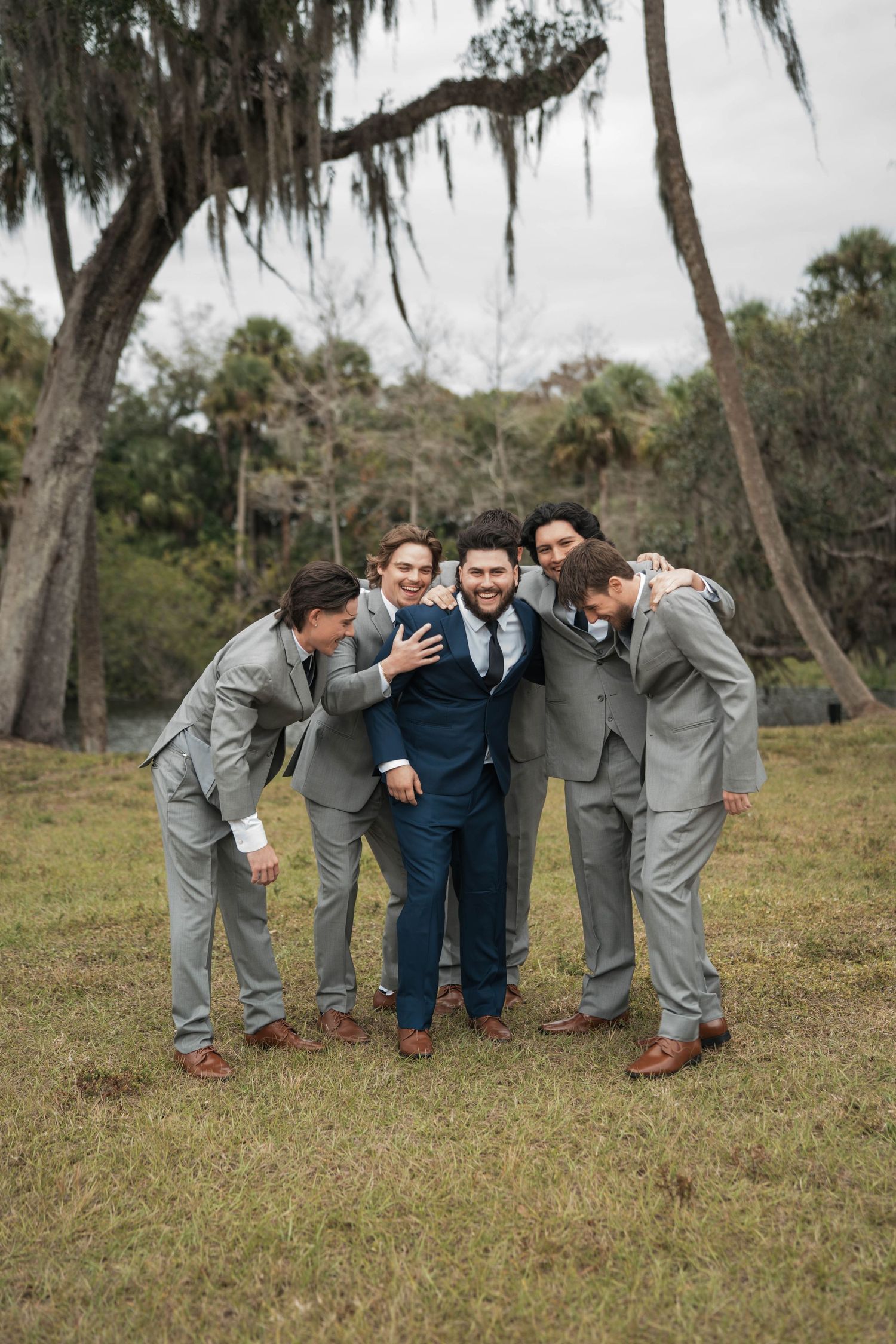 Groom in navy suit surrounded by groomsmen in gray suits outdoors under Spanish moss.