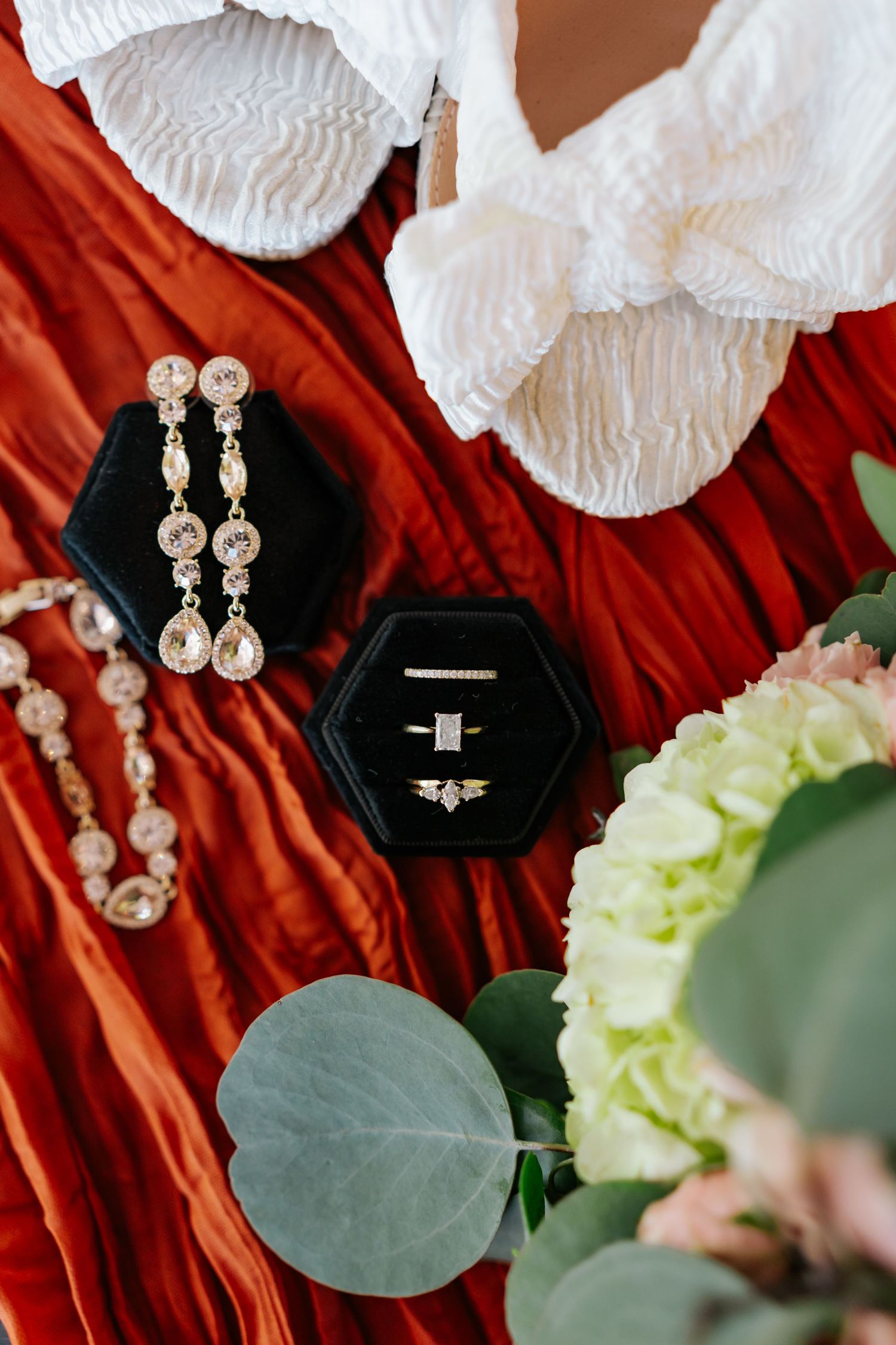 Bridal flat lay with crystal earrings, rings in black velvet box, bracelet, and hydrangea bouquet on red fabric.