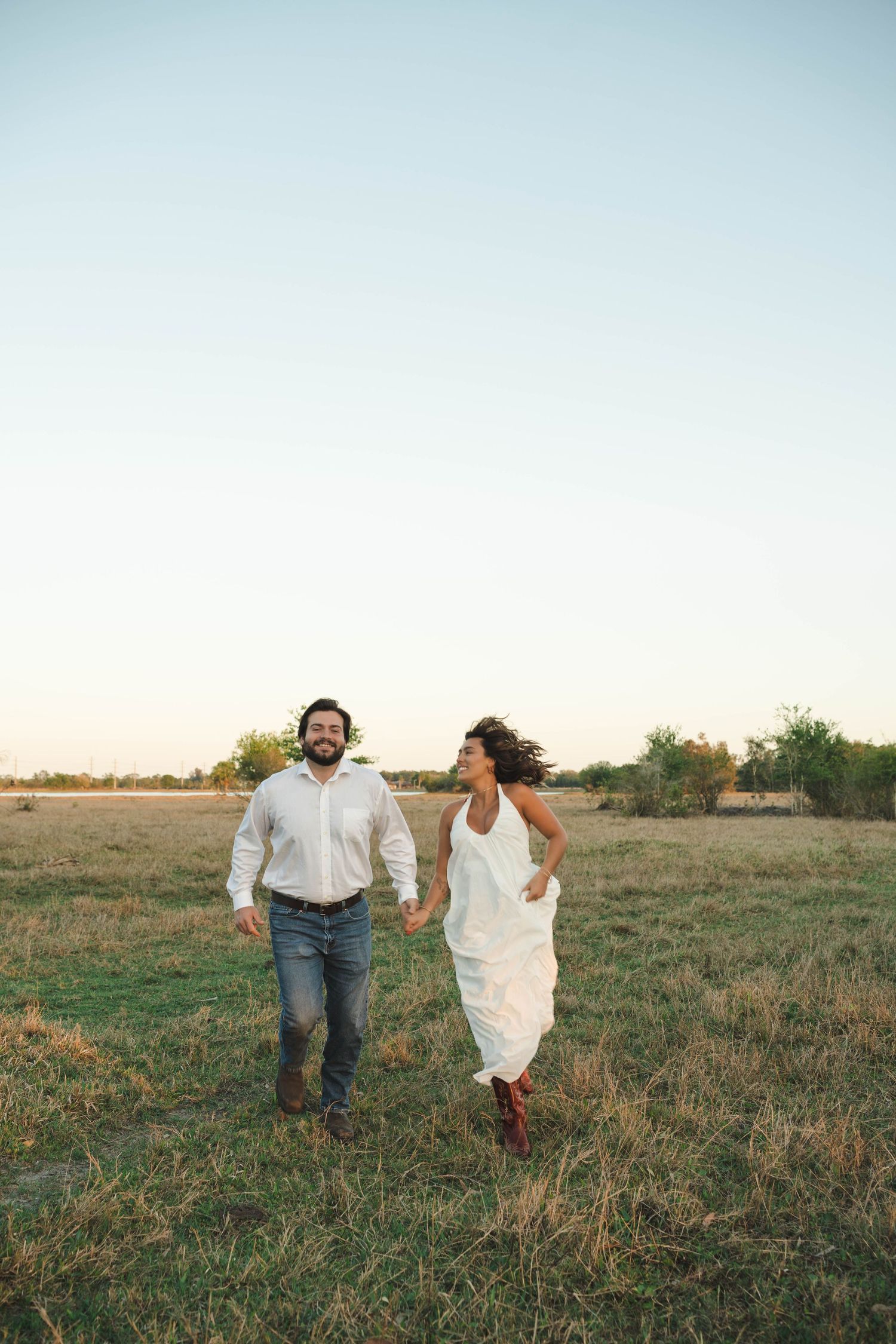 Happy couple holding hands and running through an open golden field at dusk in casual white and denim attire.