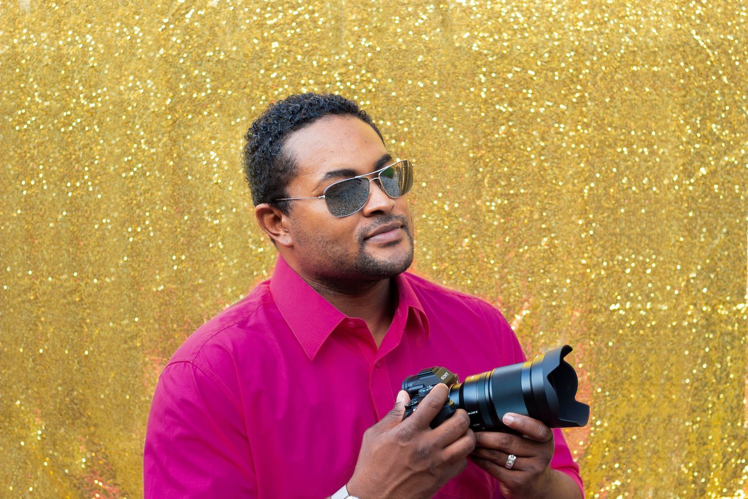 Male photographer in pink shirt holds camera against gold glitter backdrop wearing sunglasses.