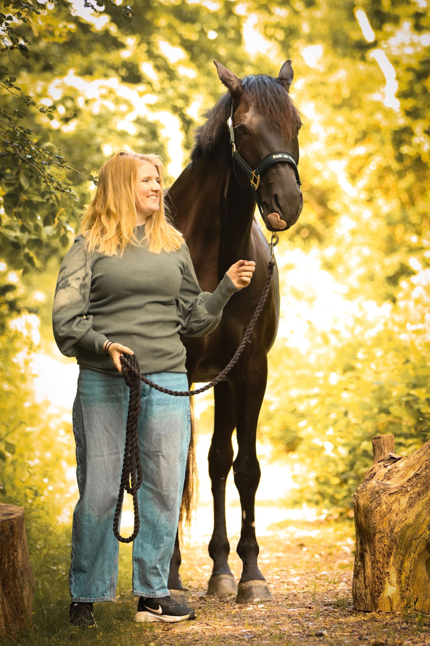 Smiling blonde woman in green sweater and jeans stands beside a dark brown horse on a sunlit forest path.