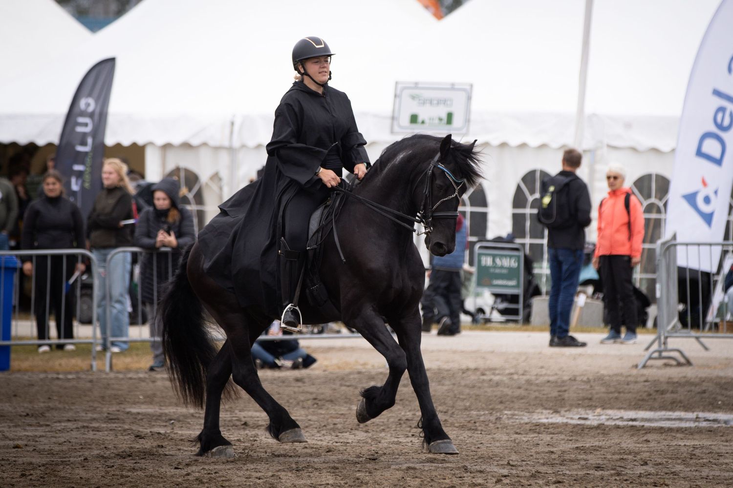 Rider in black outfit on a black horse performing at an outdoor equestrian event with spectators watching.
