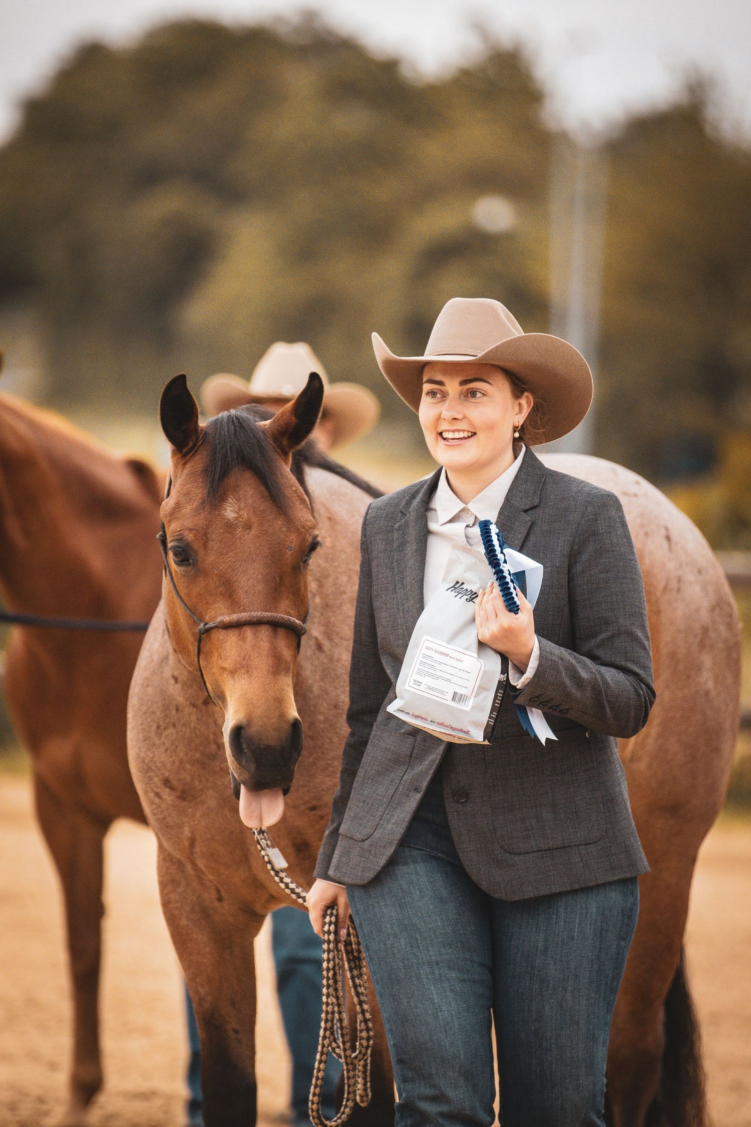 A smiling woman in a cowboy hat and gray blazer holds a lead rope beside a bay horse at an equestrian show.