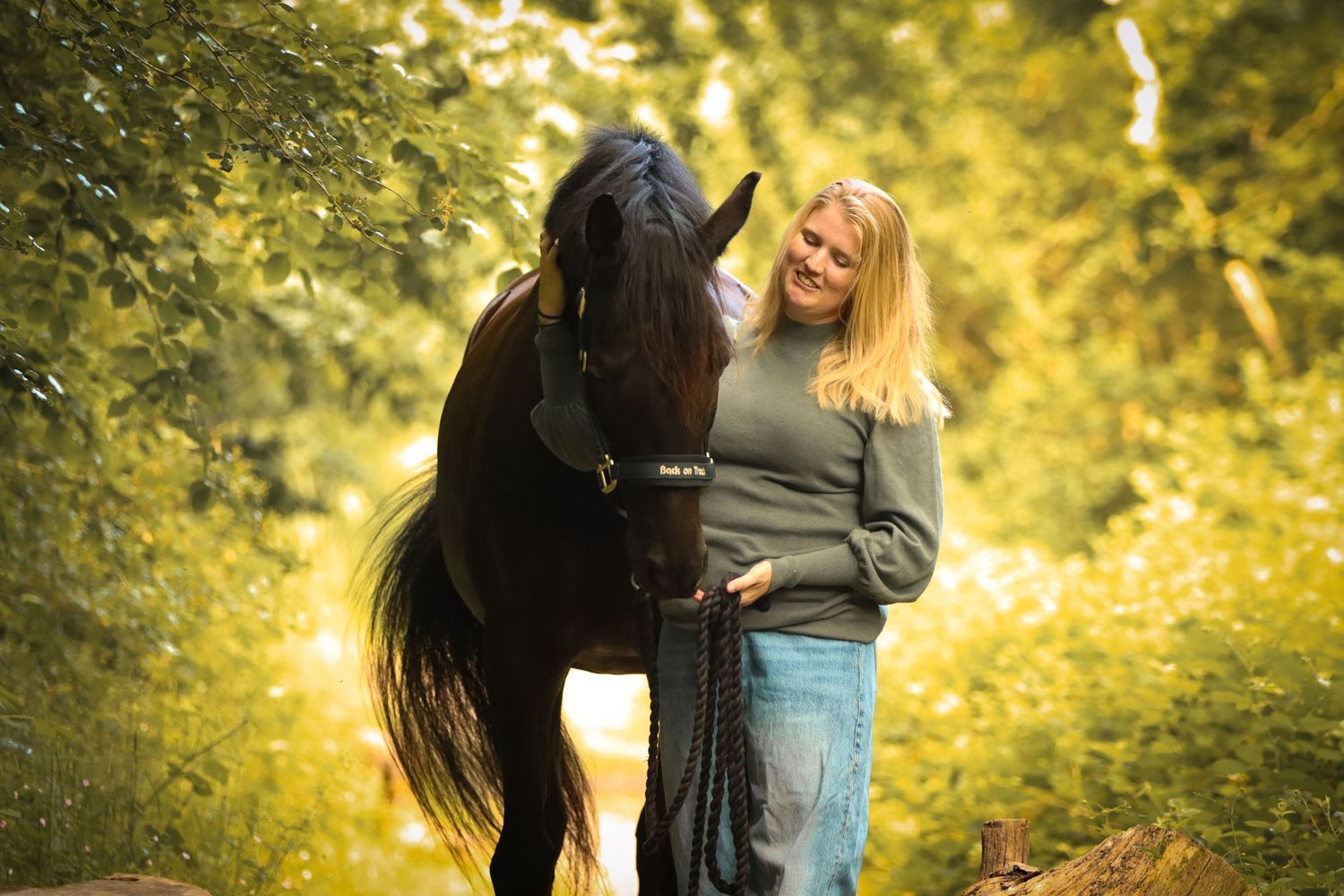 Blonde woman smiling beside a dark horse in a sunlit forest setting.