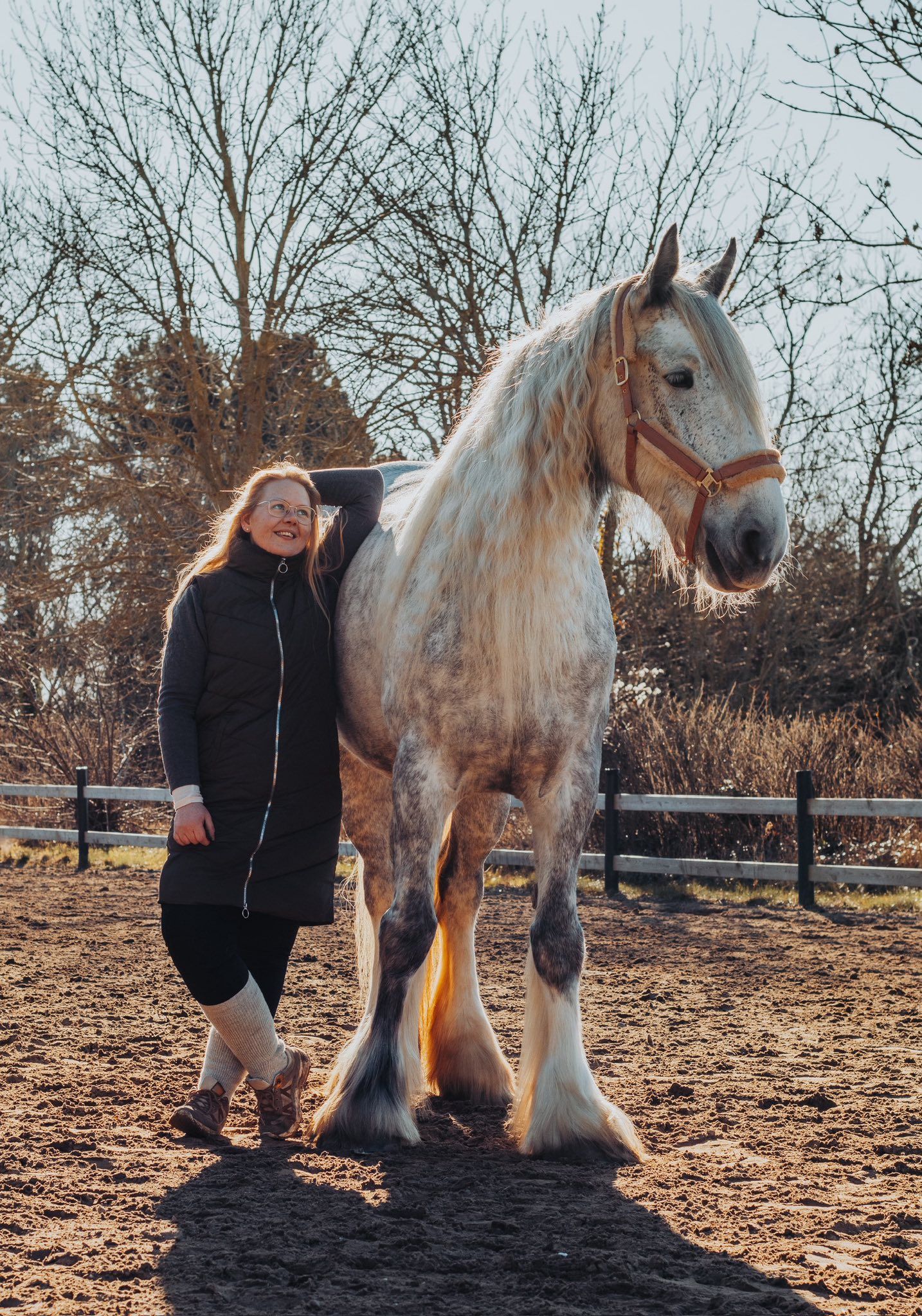 Smiling woman in black jacket posing beside a large white Gypsy Vanner horse outdoors.