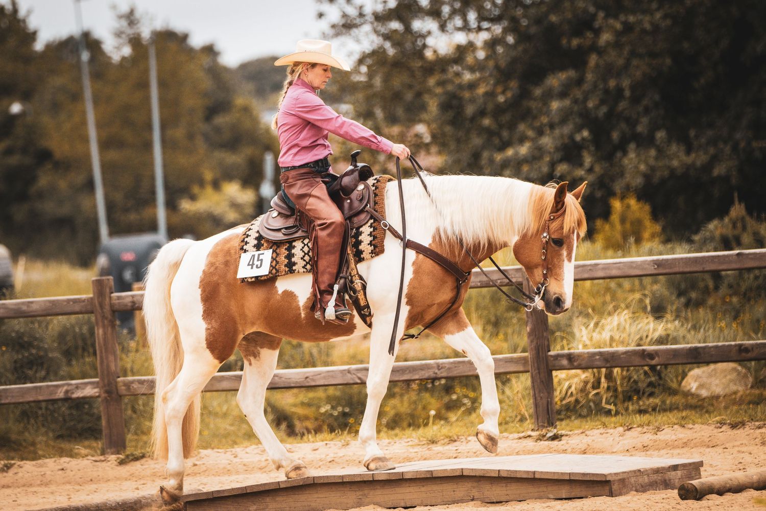 Western rider in pink shirt and cowboy hat rides a paint horse in a sandy outdoor arena.