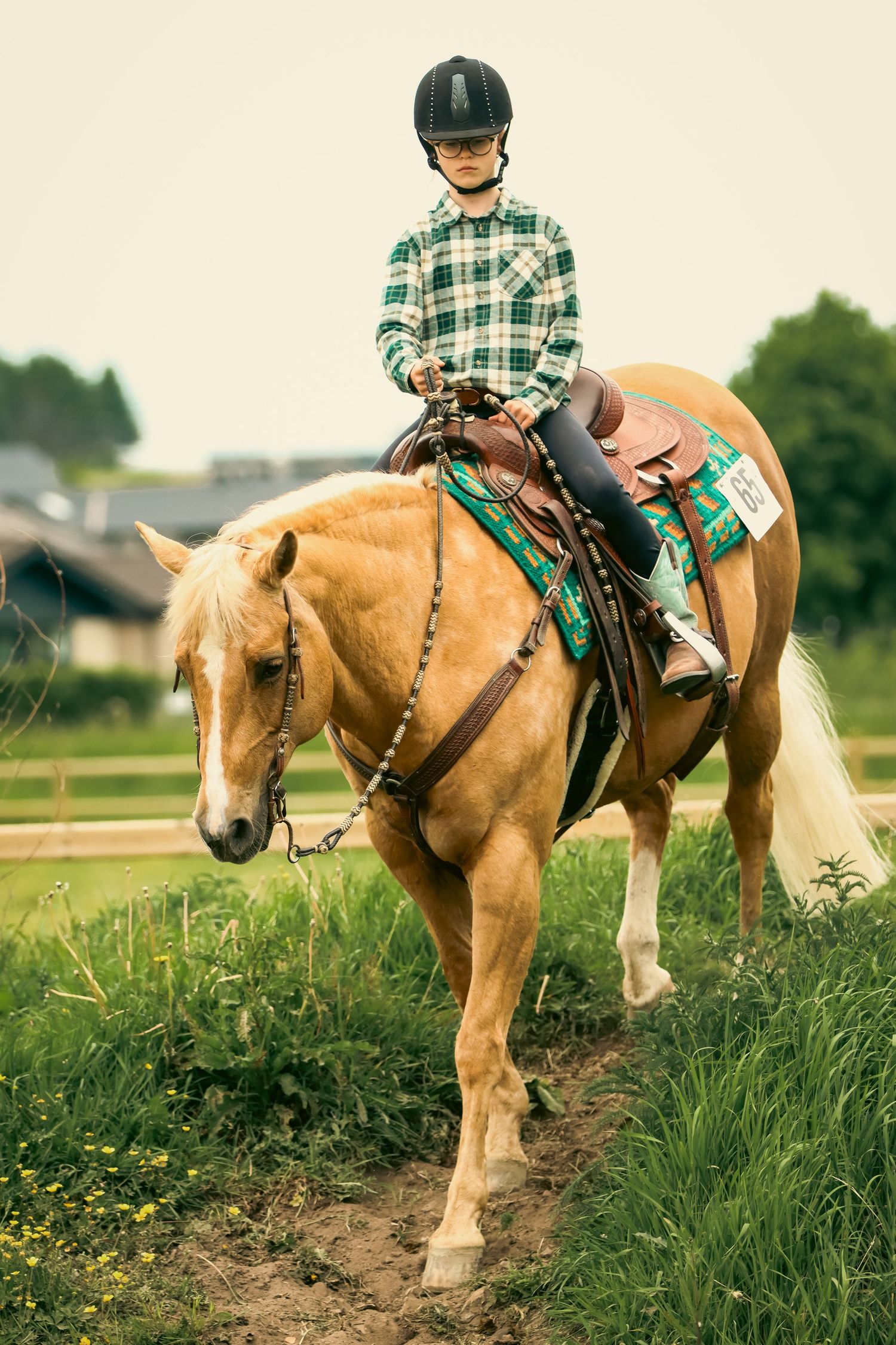 Young rider in helmet and plaid shirt on palomino horse walking through green grass field.