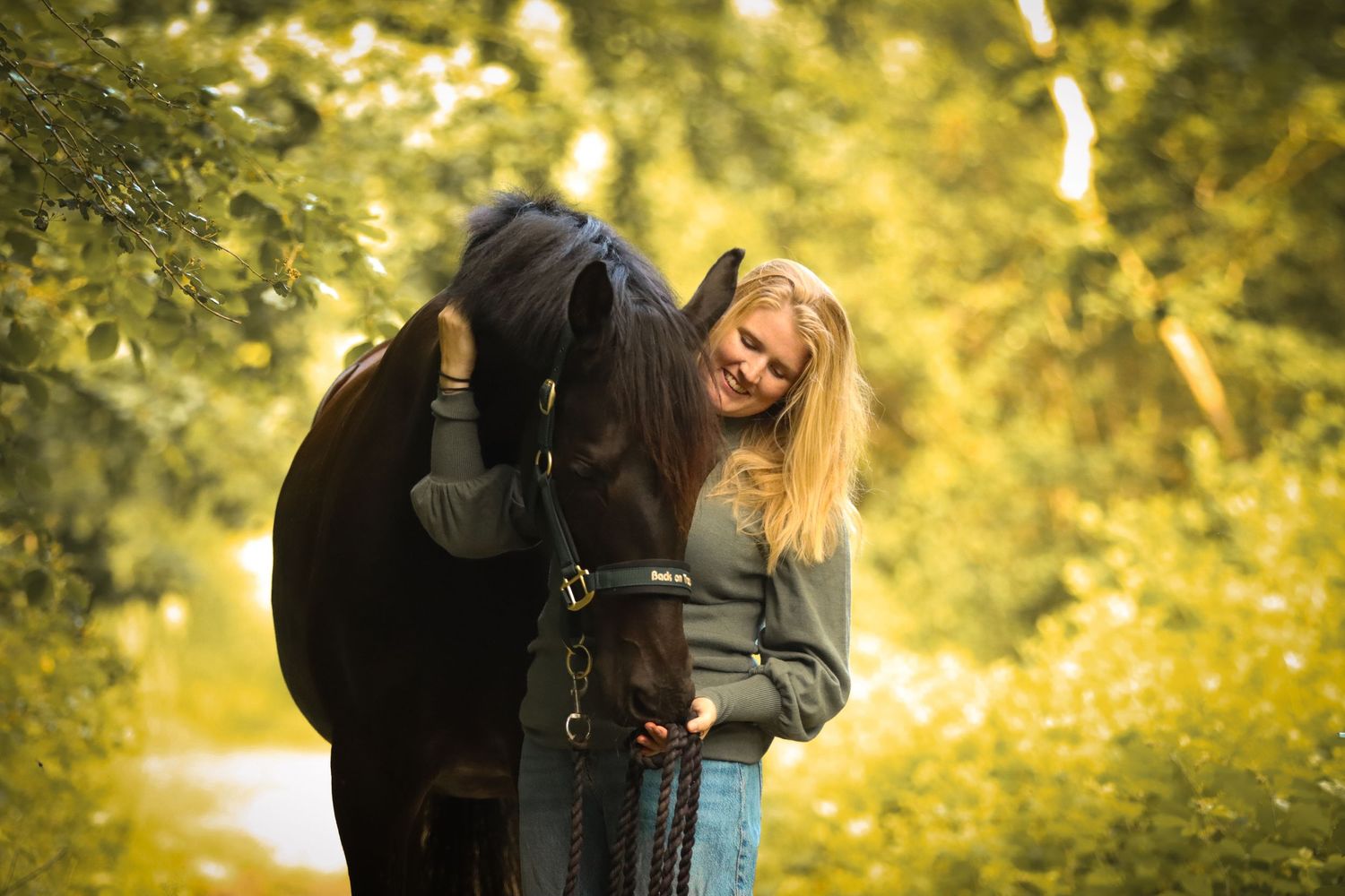 Smiling blonde woman hugging a dark horse on a sunlit forest path.
