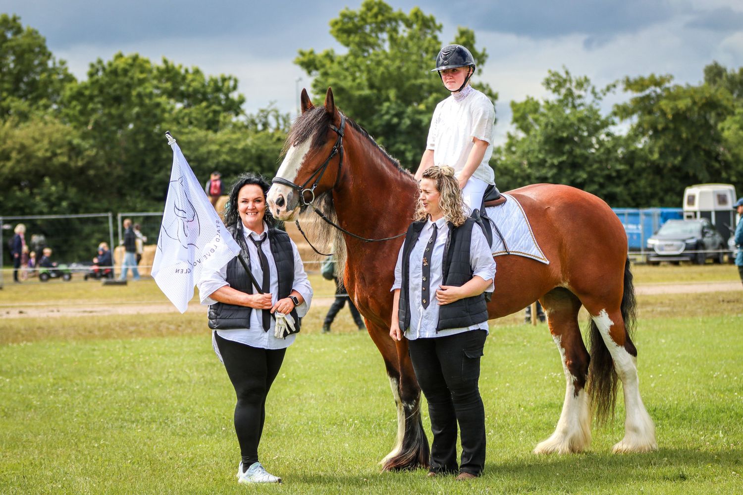 Two women in black uniforms stand with a flag beside a brown horse ridden by a child at an outdoor equestrian event.