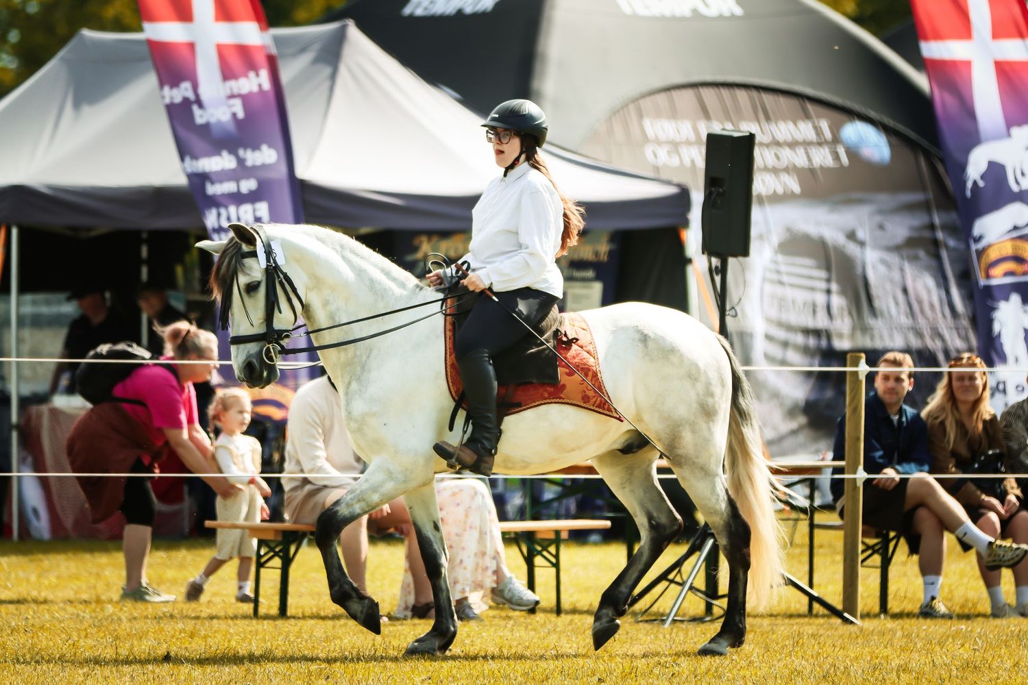 Female equestrian in white shirt riding grey horse at outdoor competition event.