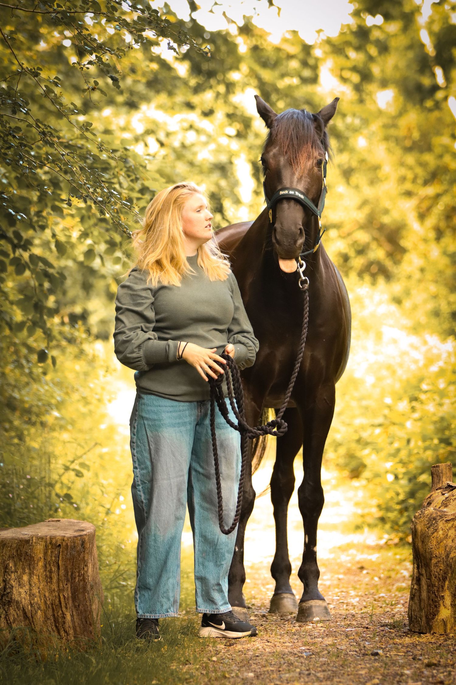 Blonde woman in green sweater and jeans stands with dark brown horse on a sunlit forest path.