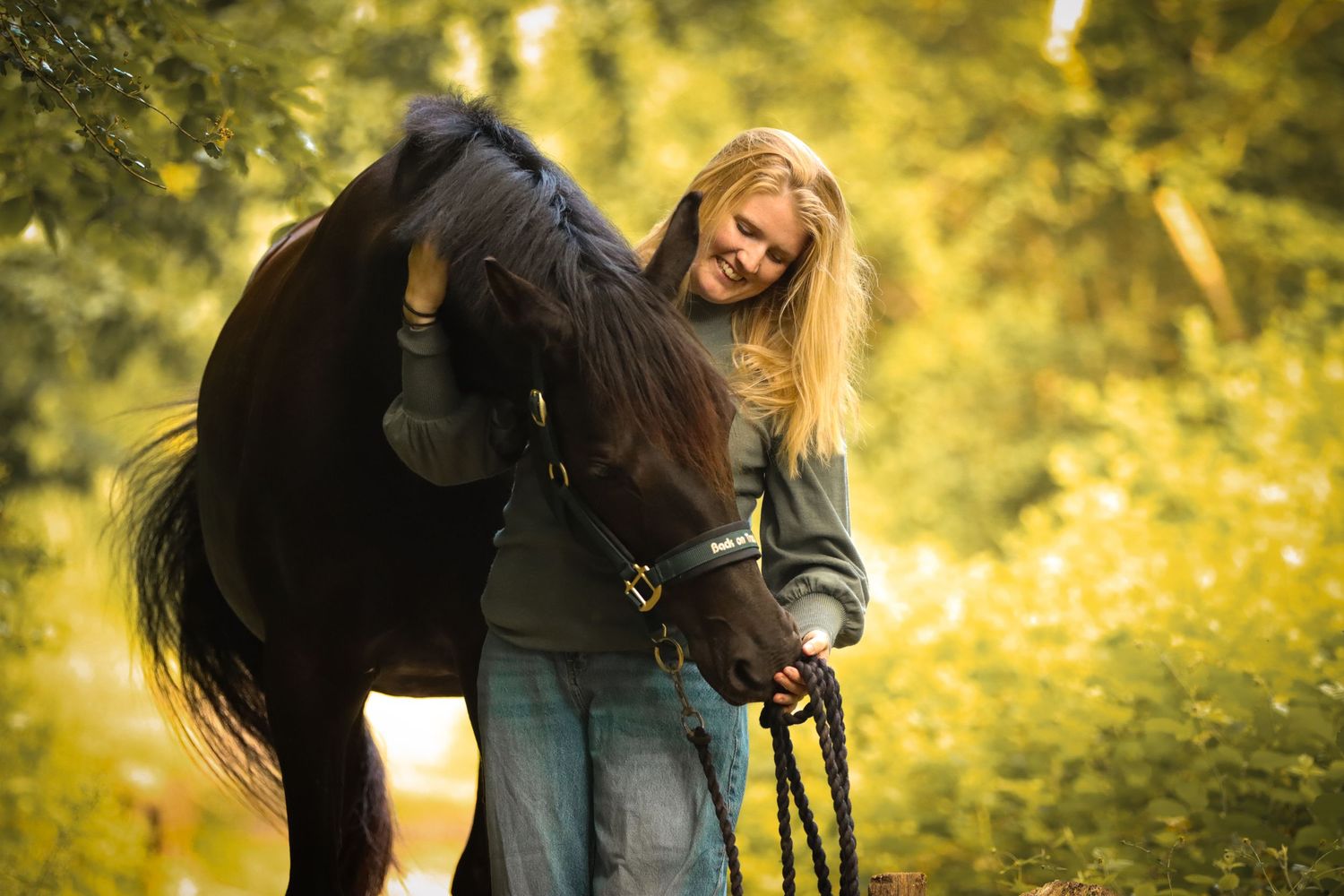Blonde woman smiling while affectionately hugging a dark horse outdoors in a sunlit green forest setting.