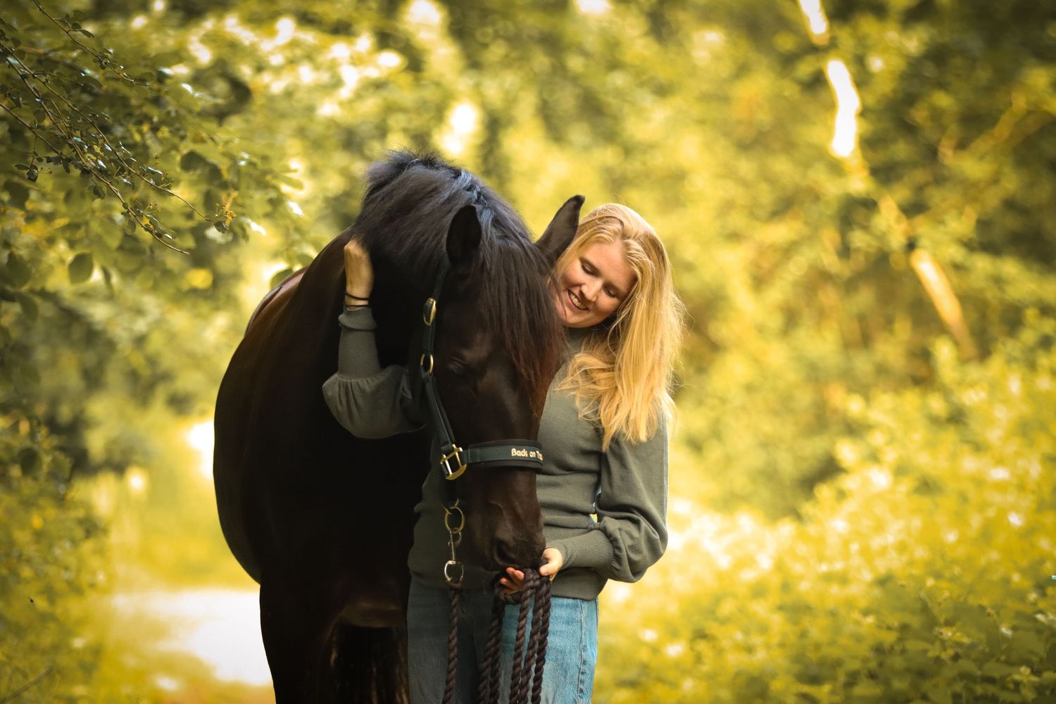 Smiling blonde woman hugging a dark brown horse on a sunny forest path surrounded by lush green trees.