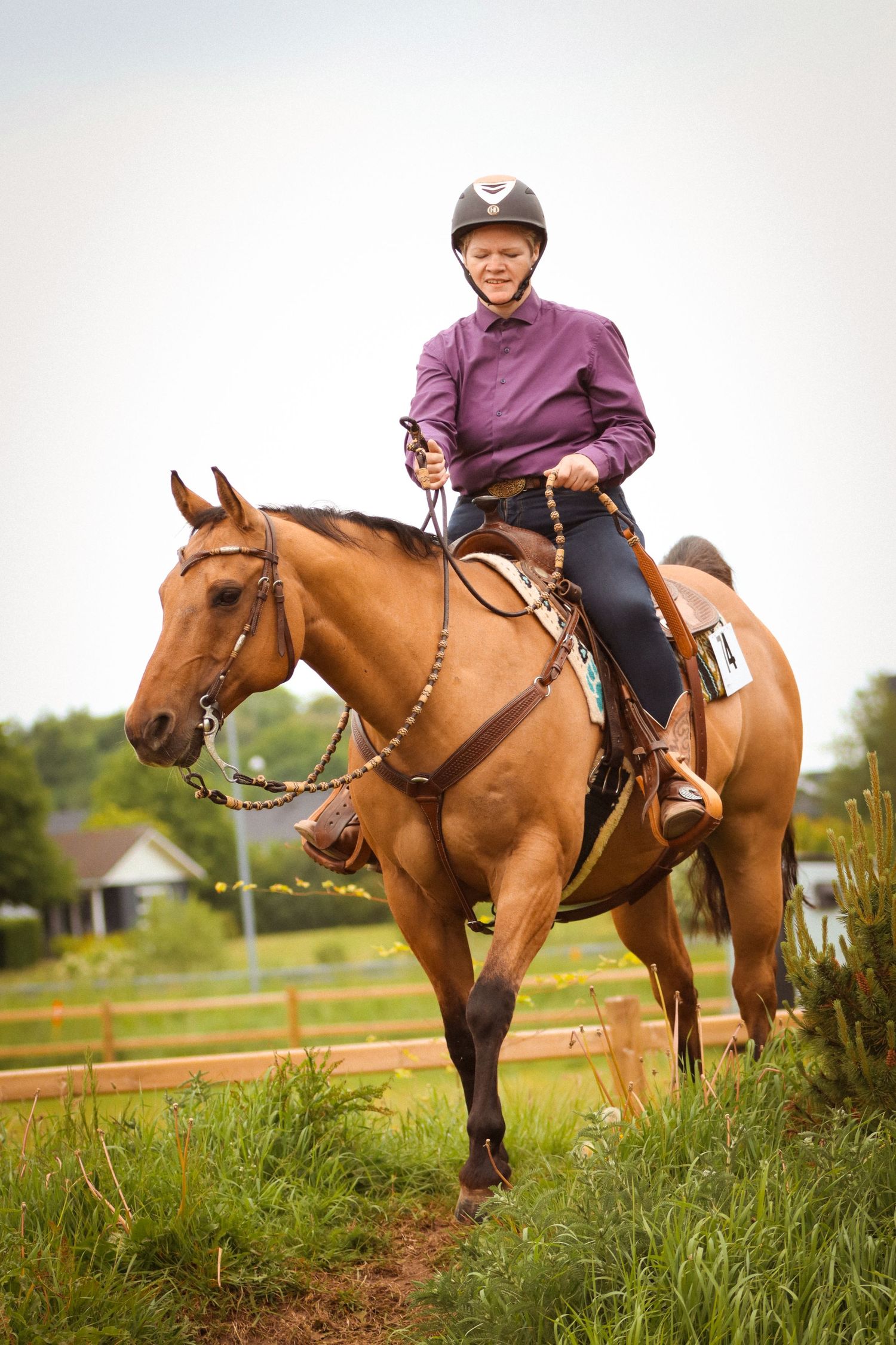 Smiling young rider in purple shirt and helmet rides a chestnut horse through a grassy outdoor trail.