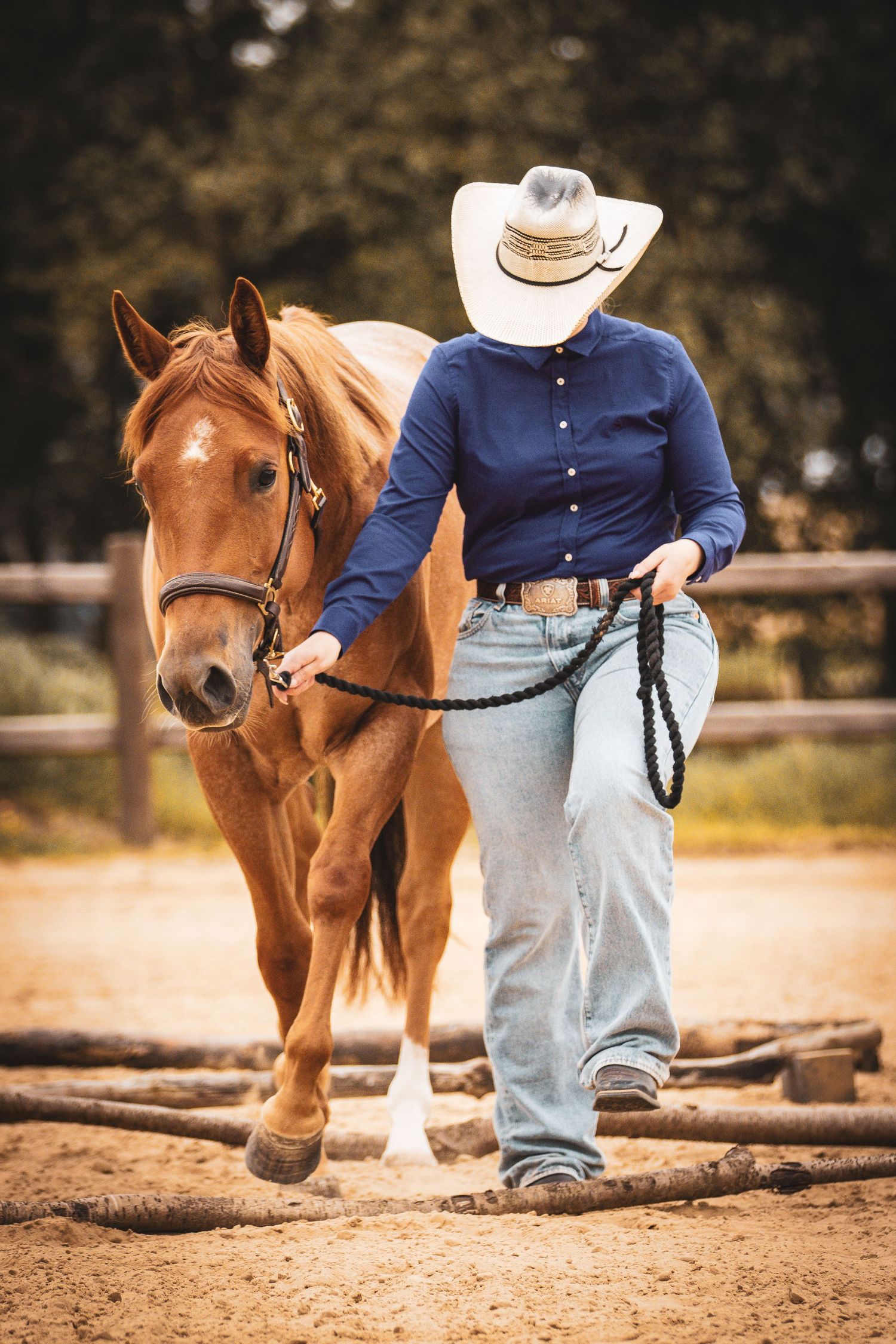 Cowboy hat-wearing rider leads a chestnut horse by a black lead rope in a sandy paddock.